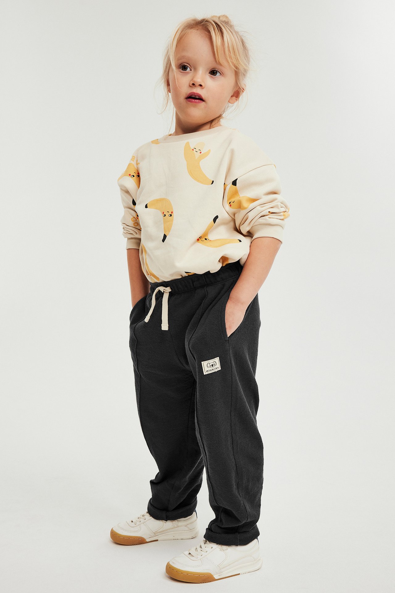 Framifrån: Ett barn i svarta mjukisbyxor med dragsko, sidofickor och en tröja med banantryck.