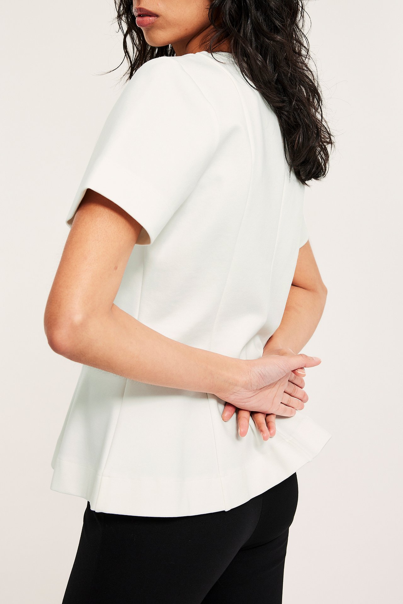 Sidebilde av kvinne i en offwhite kortermet peplumtopp og svarte bukser, med hendene knyttet bak ryggen.