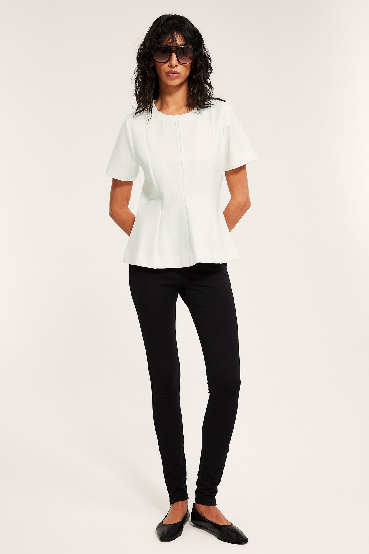 Front: Kvinne med offwhite kortarmet peplumtopp, svarte smale bukser, svarte ballerinasko og mørke solbriller.
