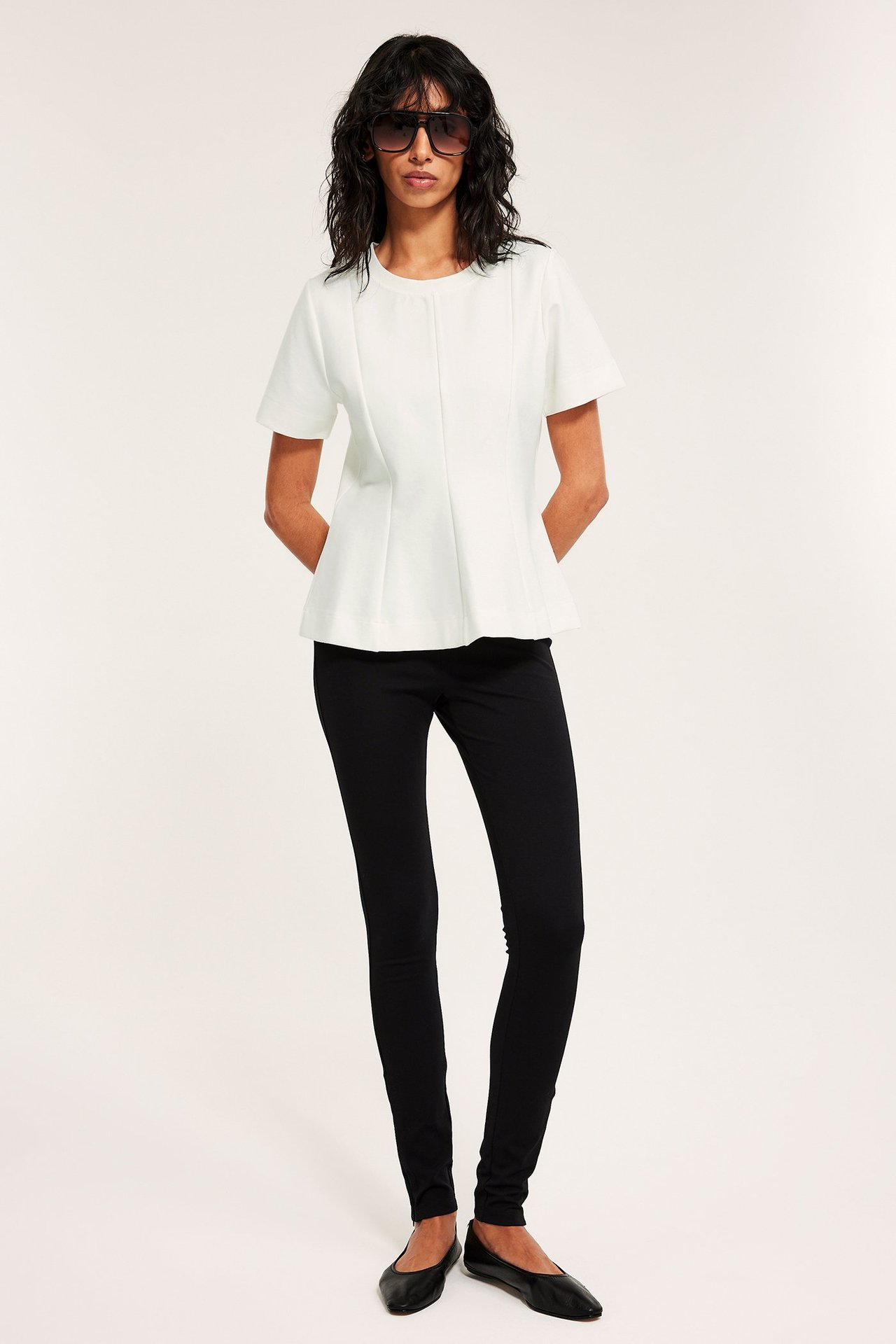 Framsida: Kvinna i offwhite kortärmad peplumtopp, svarta smala byxor, svarta ballerinaskor och mörka solglasögon.