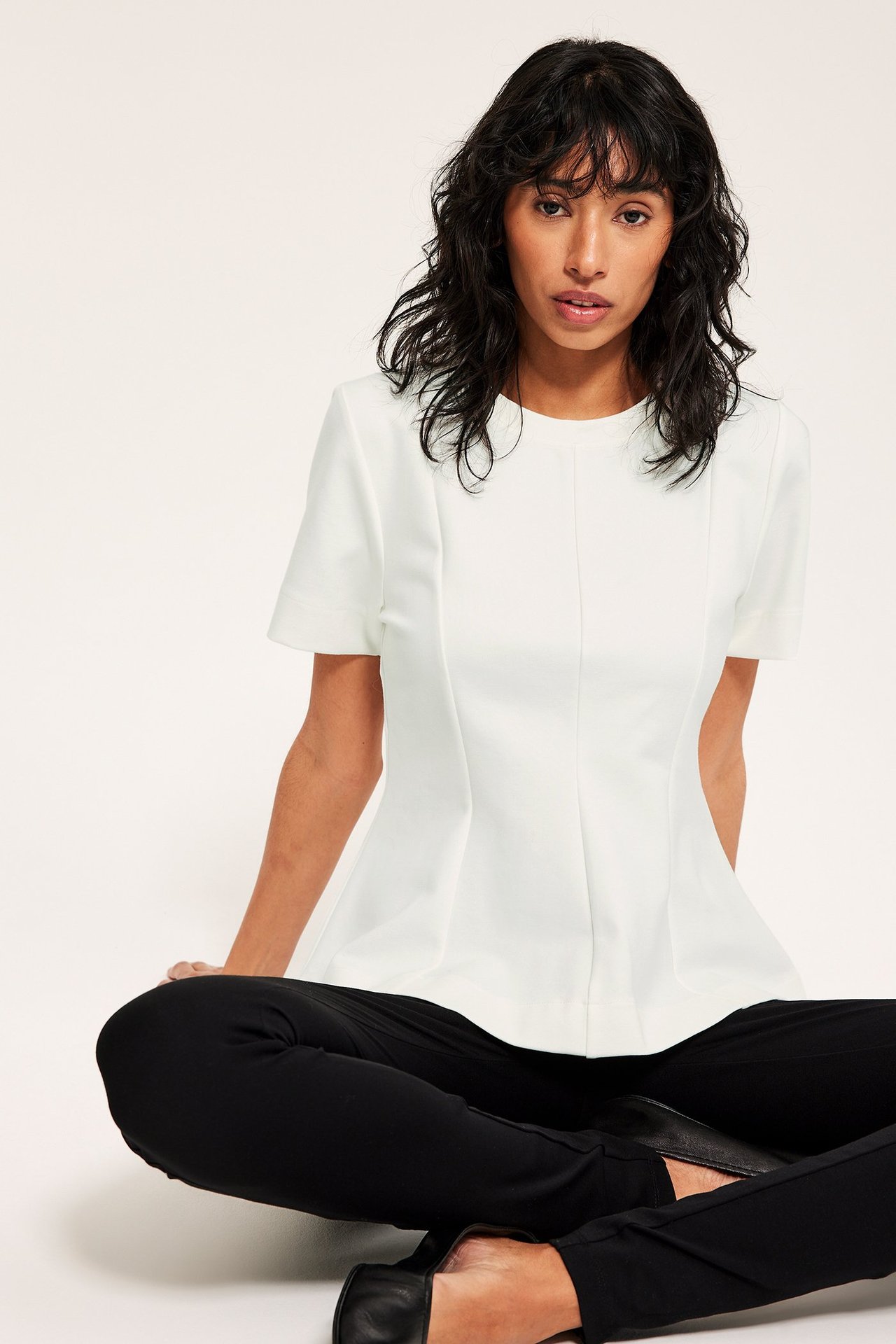 Widok z przodu: kobieta w kremowym krótkim topie peplum, czarnych spodniach i czarnych płaskich butach.