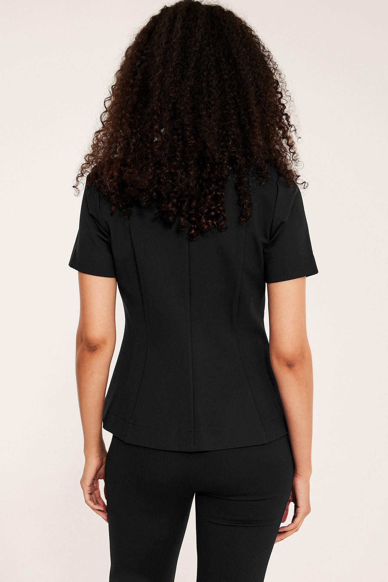 Bakfra av en kvinne i svart kortermet peplumtopp med utsvingt skjøt og svarte bukser.