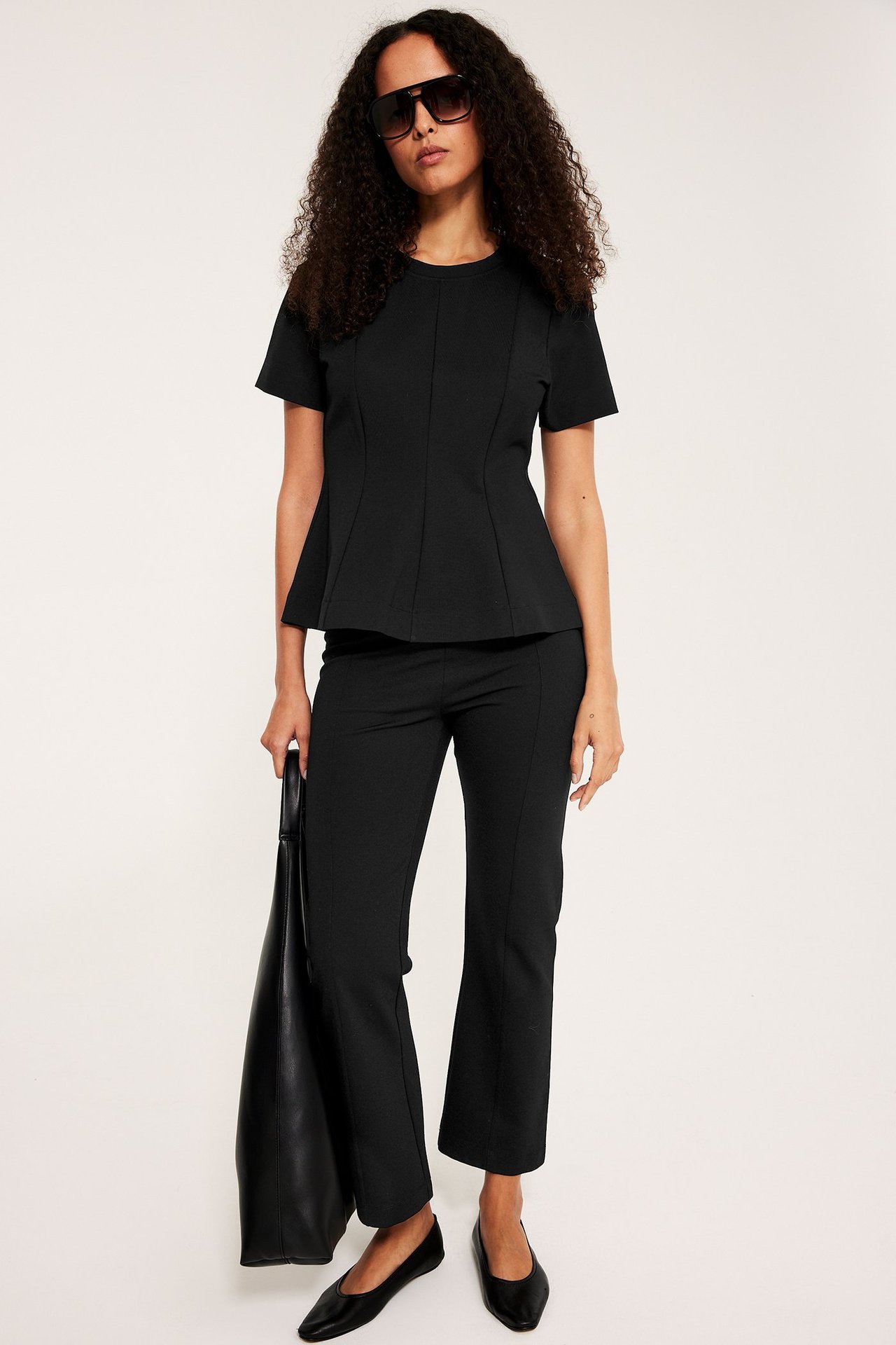 Framsida: Kvinna i svart kortärmad peplumtopp, svarta byxor, platta skor, solglasögon, håller en svart tygväska.