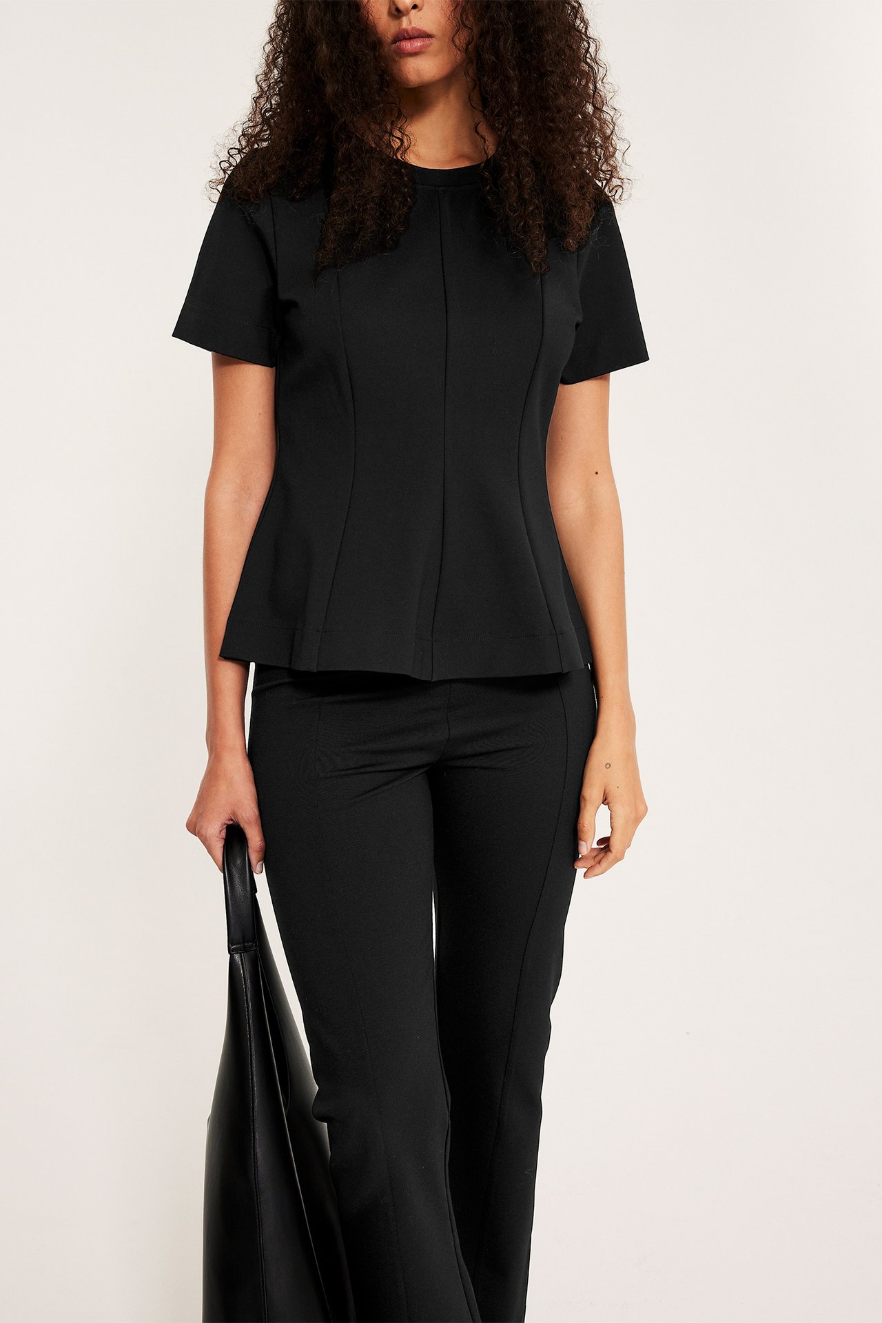 Forfra: Kvinne i svart kortermet peplumtopp, stylet med svarte bukser, holder en svart håndveske.