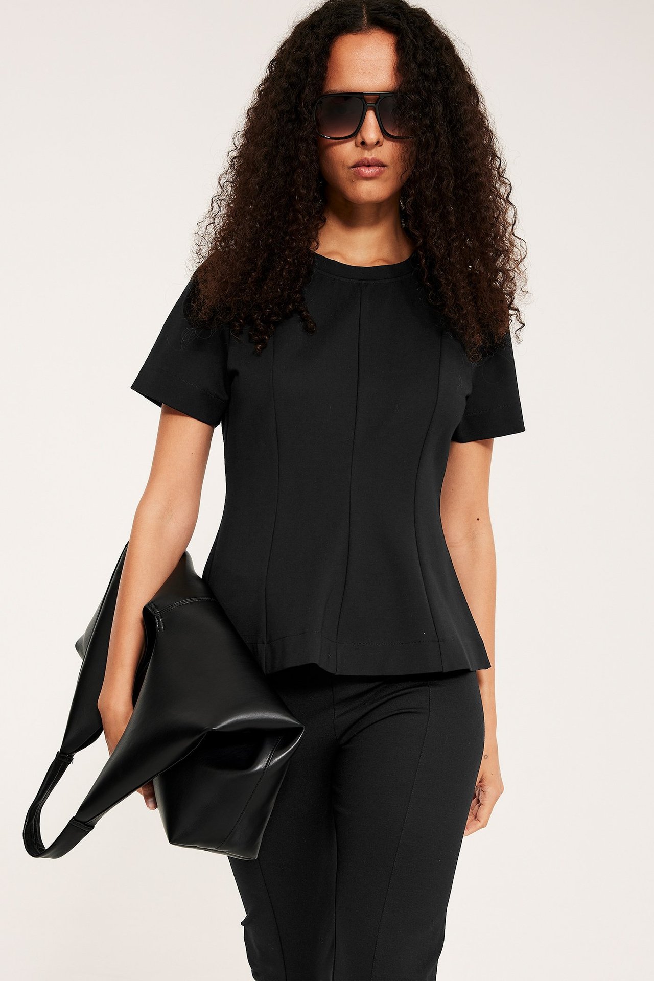 Kvinna i svart kortärmad peplumtopp, svarta byxor, solglasögon, håller en svart handväska.