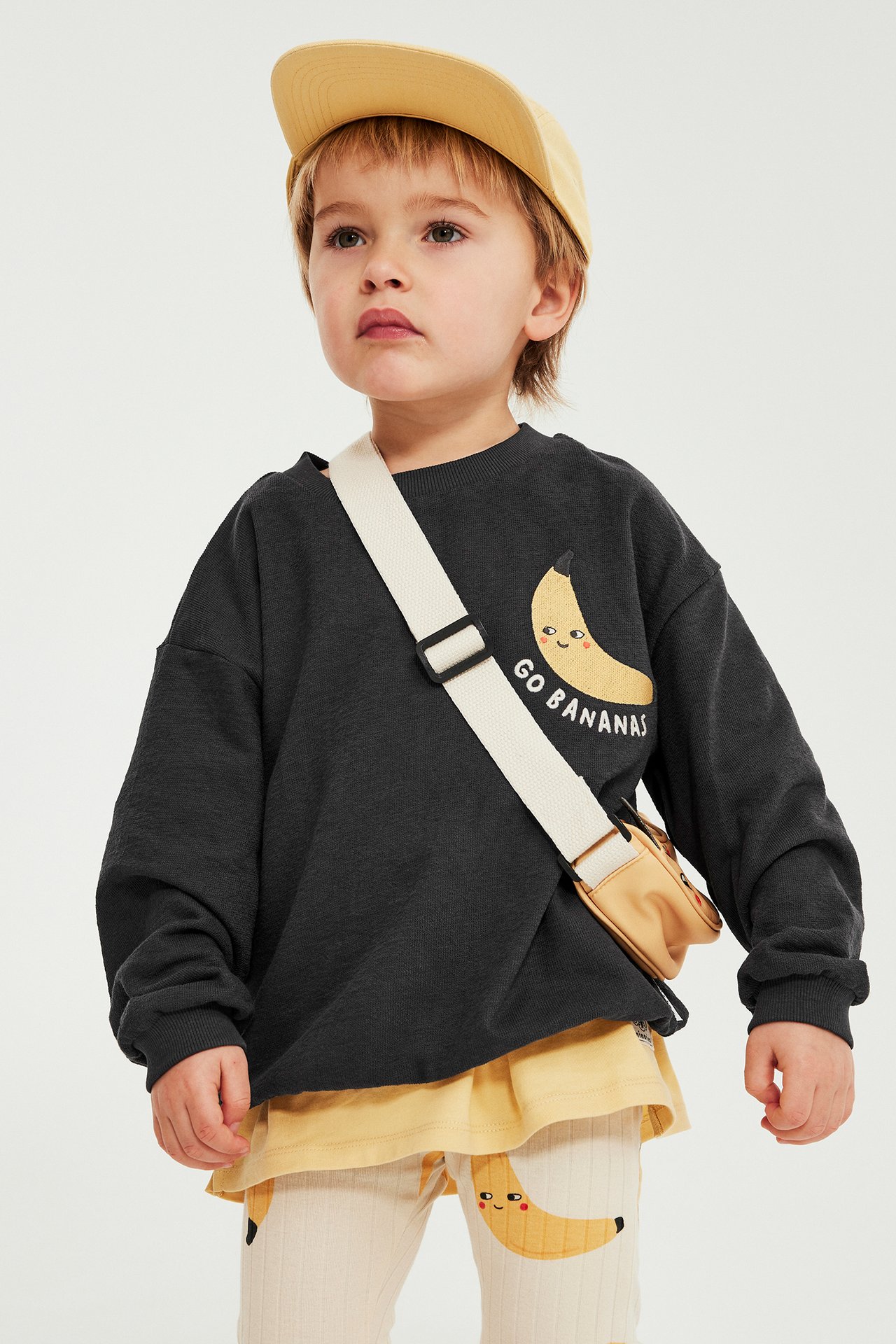 Framsida: Ett barn i svart bomullssweatshirt med bananbrodyr, beige keps, och ribbade bananleggings.