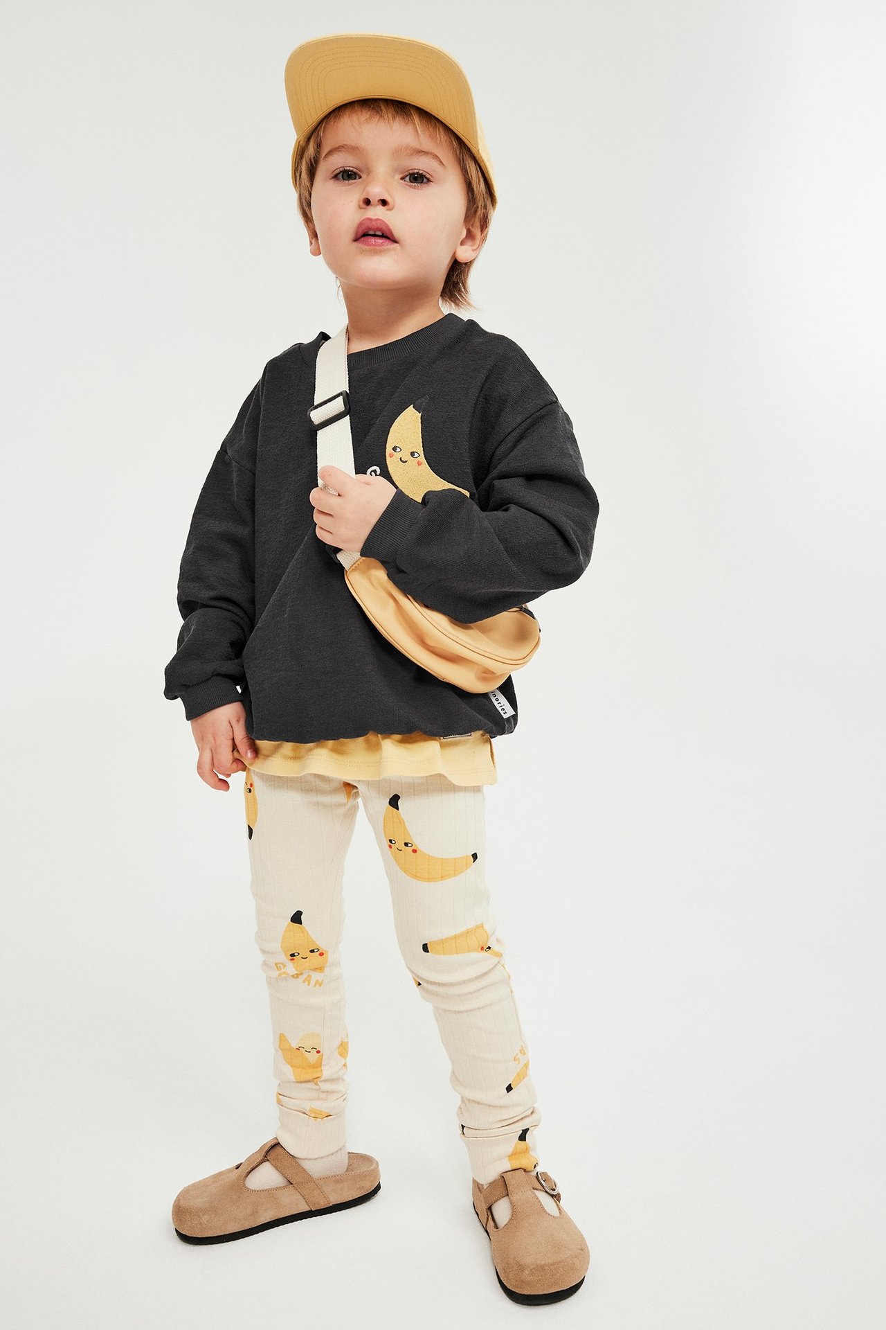 Barn bär svart sweatshirt med bananbrodyr, beige bananleggings, gul t-shirt, senapsgul keps, beige midjeväska och bruna tofflor.