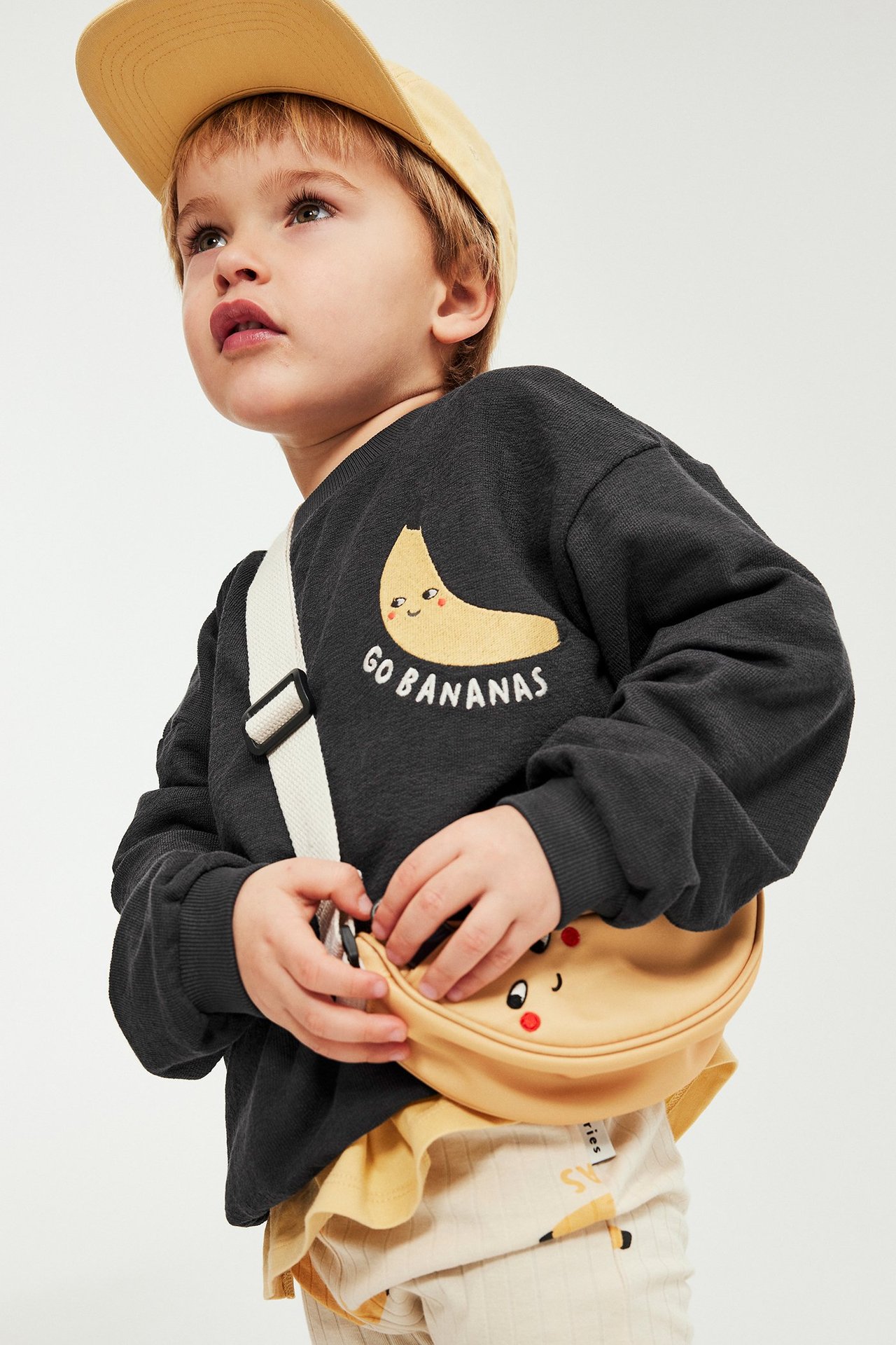 Edestä: lapsi mustassa bananikuviollisessa collegepaidassa, keltainen lippalakki, keltainen vyölaukku ja vaaleat ribbihousut.
