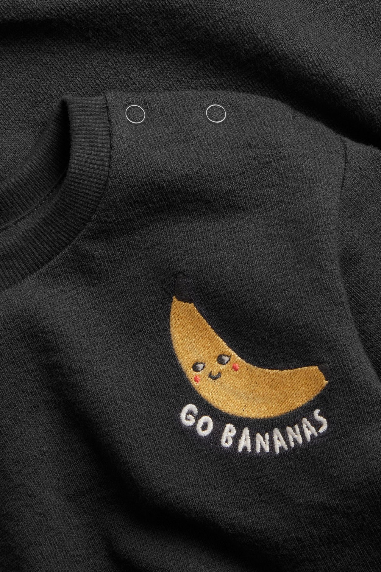 Lähikuva mustasta puuvillaisesta vauvan collegepaidasta banaanikirjailulla, 'GO BANANAS' -tekstillä ja olkanapeilla.