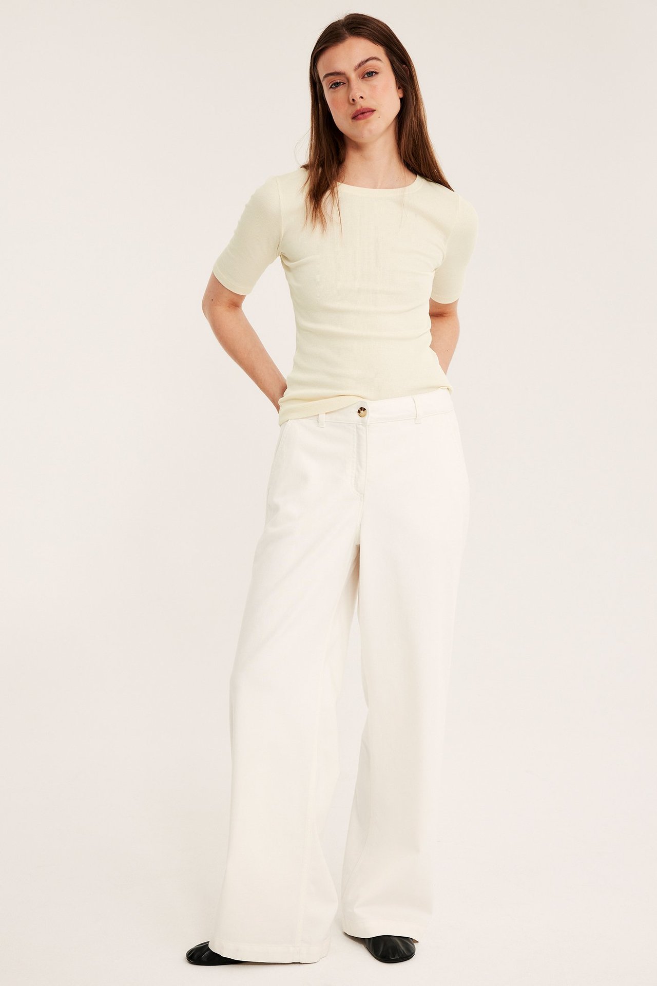Frontbilde av kvinne i en offwhite ribbet kortermet topp, offwhite vide bukser og svarte flate sko.