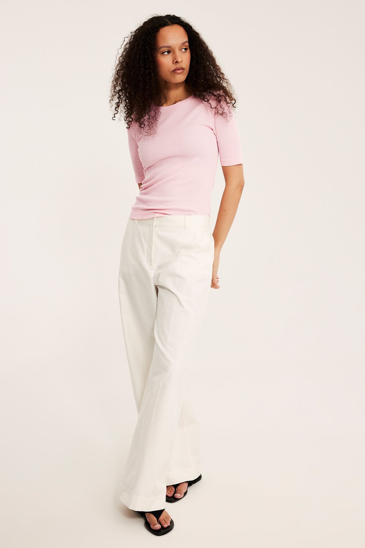 Widok z przodu: kobieta w różowym prążkowanym topie, białych szerokich spodniach i czarnych klapkach.