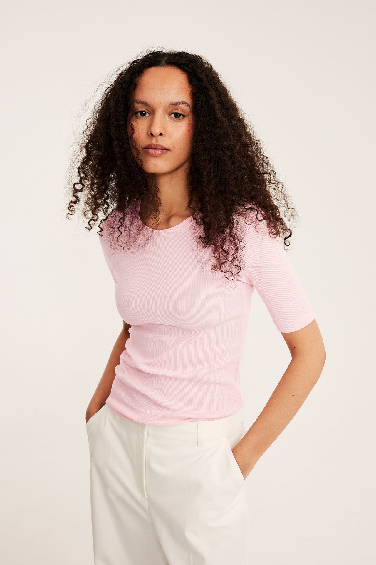 Framifrån, kvinna i rosa ribbad kortärmad topp och vita byxor med händerna i fickorna.