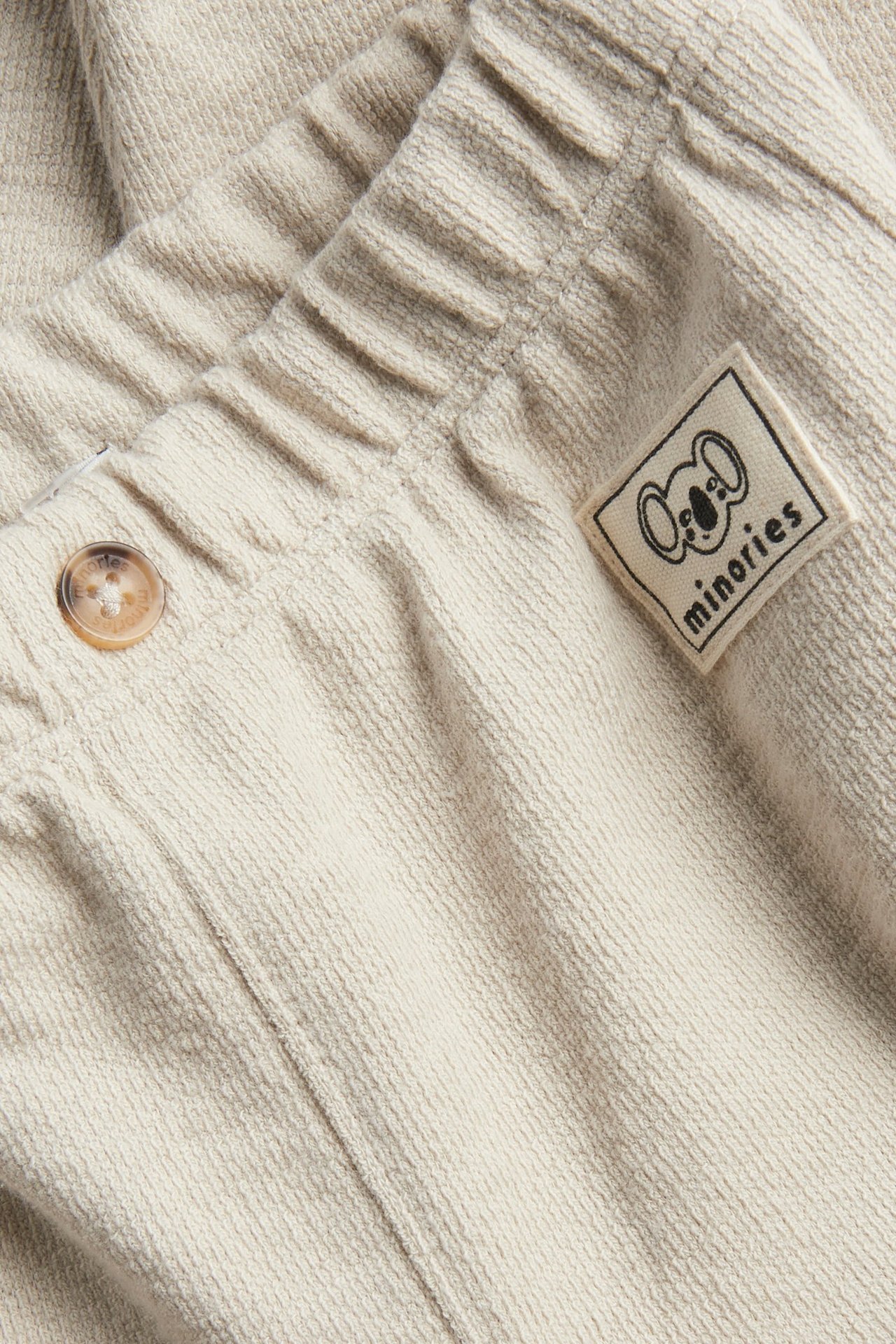 Lähikuva beigestä yksivärisestä puuvillaisesta vauvan collegehoususta, jossa joustava vyötärö ja koristenappi.