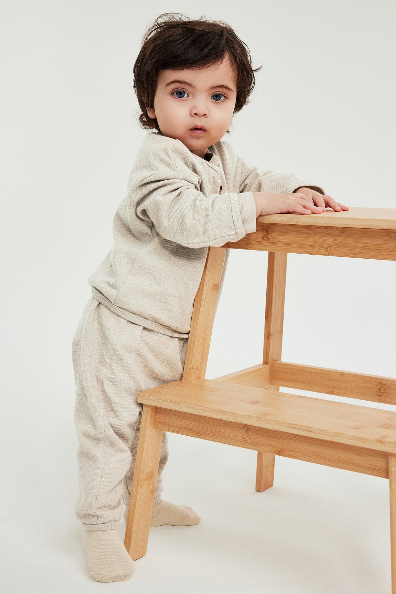 Edestäpäin: Vauva käyttää beigejä puuvillahousuja ja yhteensopivaa pitkähihaista paitaa, pitelee jakkaraa.