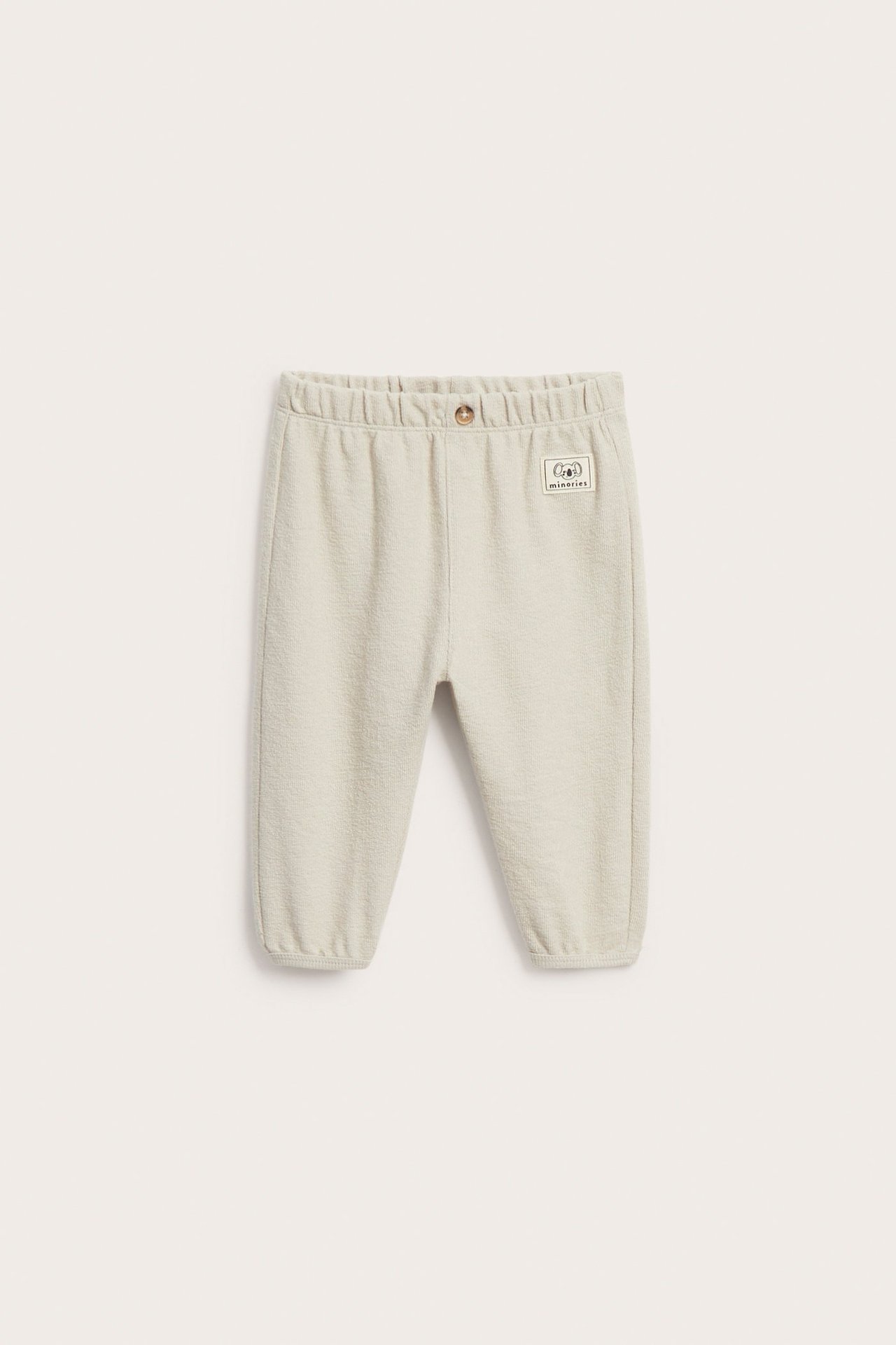 Etunäkymä yksivärisistä beigenvärisistä puuvillavauvan collegehousuista joustovyötäröllä, valenapilla ja resorimanseteilla.