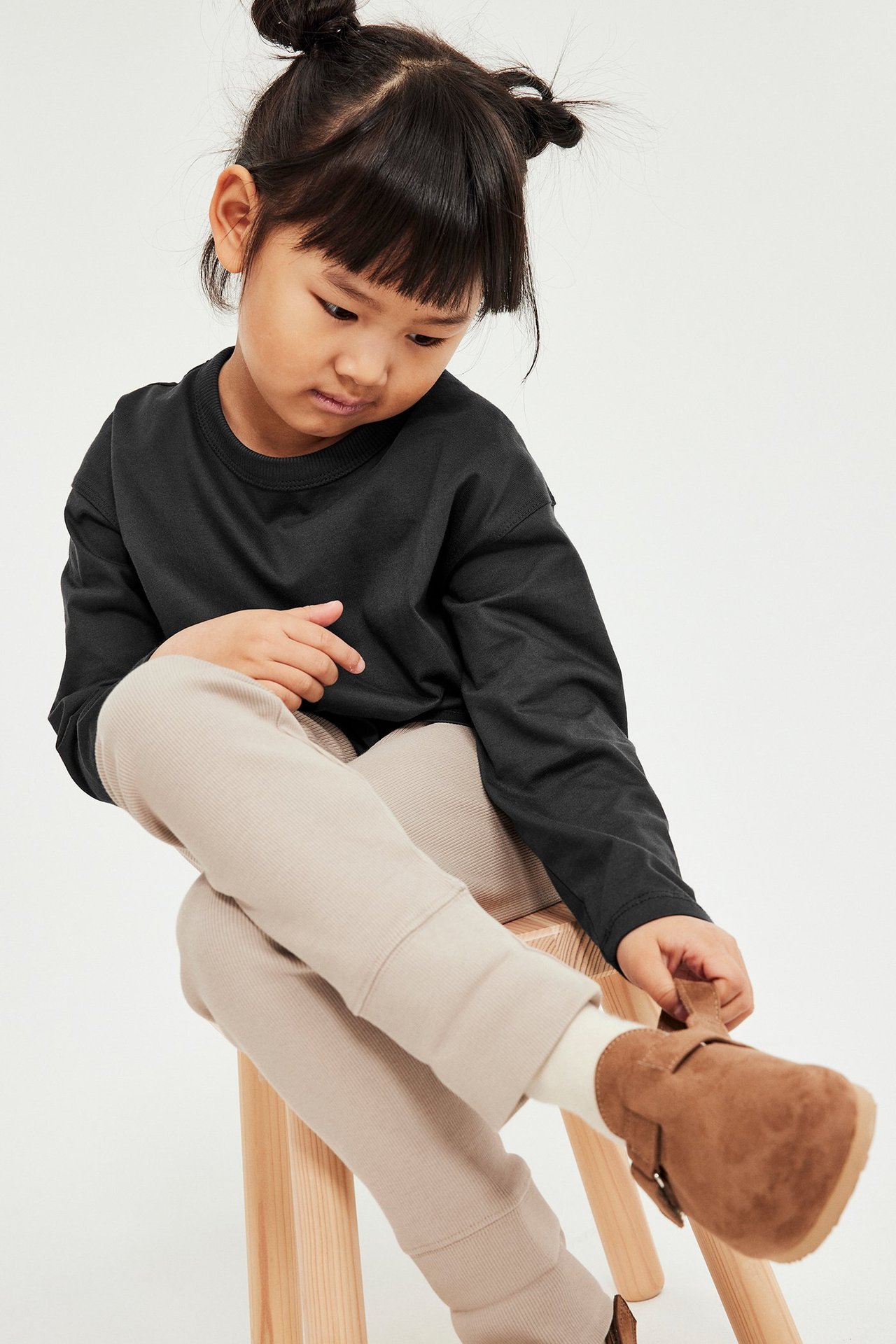 Frontowe ujęcie dziecka: czarny bawełniany t-shirt z długim rękawem, beżowe legginsy, brązowe buty.