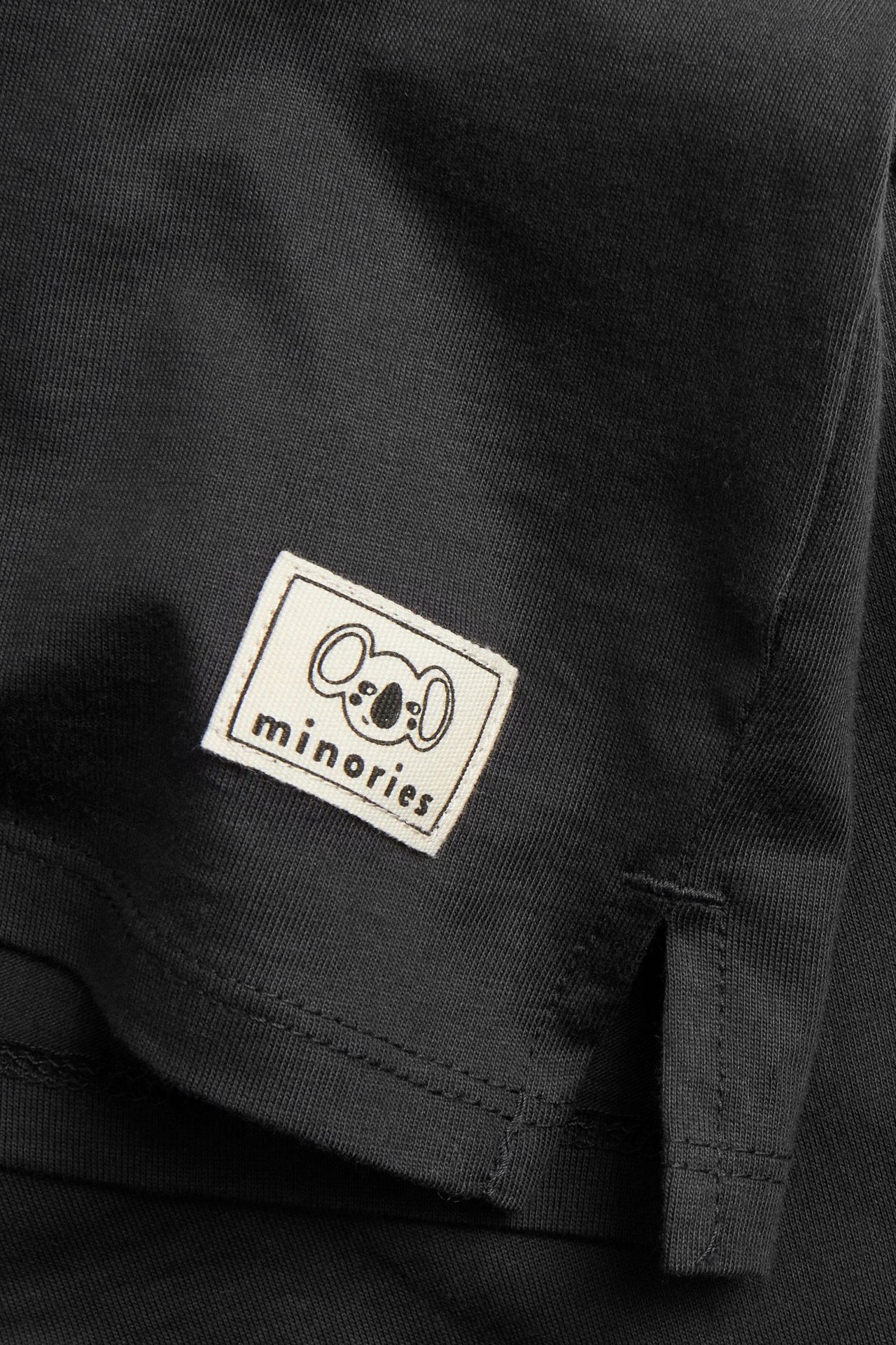 Zbliżenie na czarny bawełniany T-shirt z długim rękawem, z białą naszywką 'minories' koali i rozcięciem z boku.