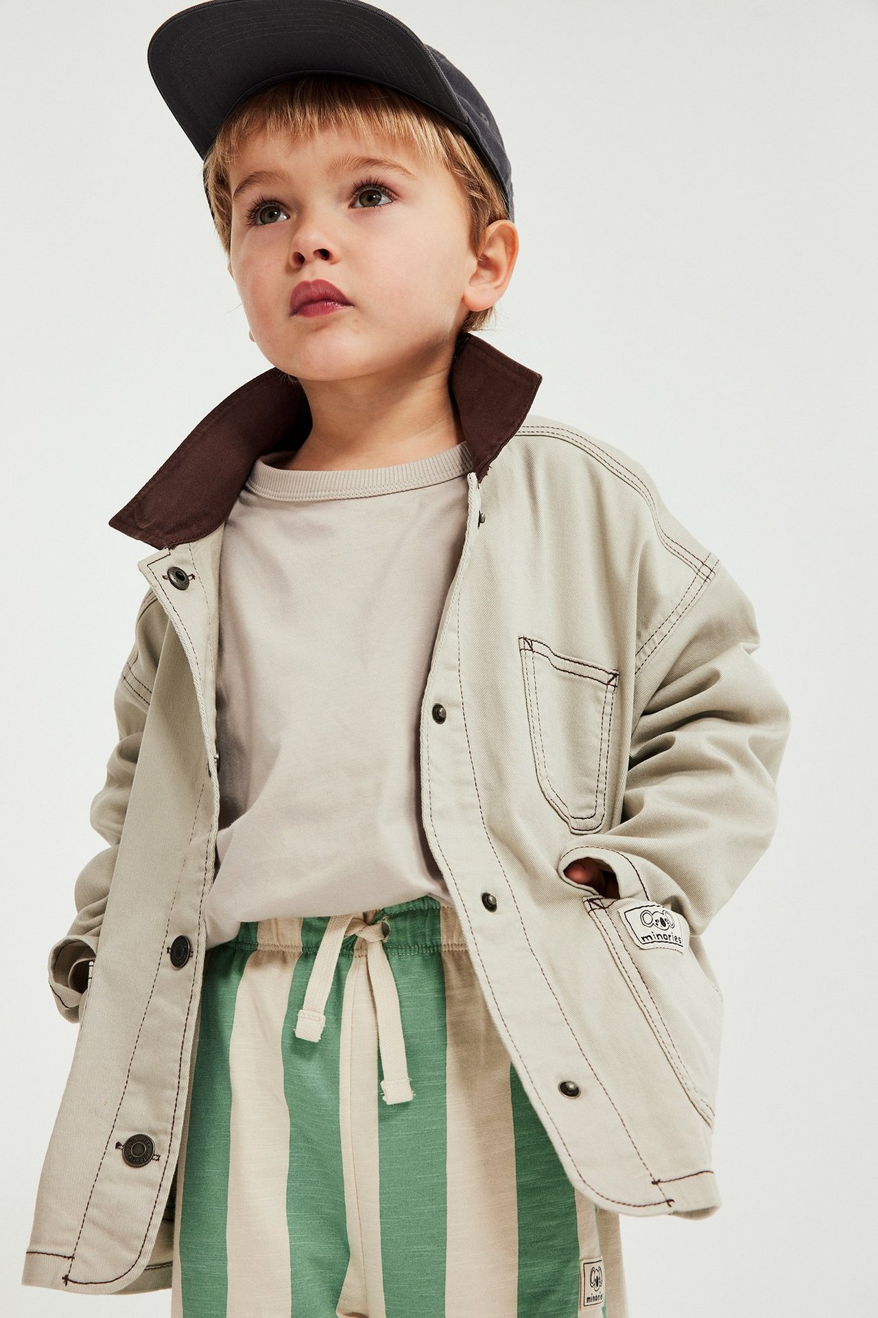 Widok z przodu dziecka w beżowym bawełnianym t-shircie, jasnej kurtce, pasiastych spodniach i ciemnej czapce.