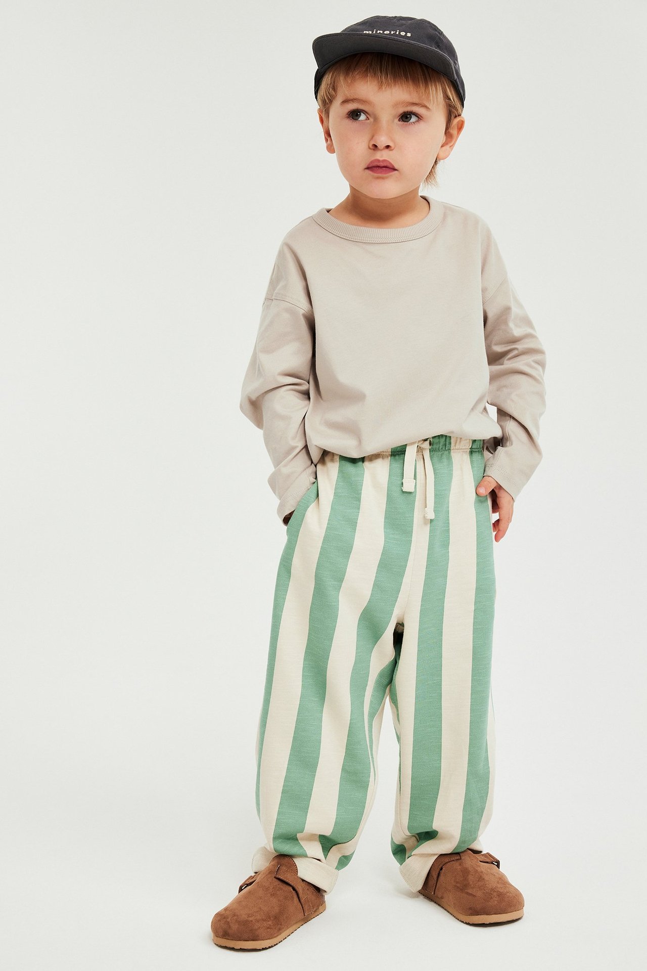 Frontbilde av et barn i en beige langermet t-skjorte, grønn- og hvitstripete joggebukse, mørkegrå caps og brune tøfler.