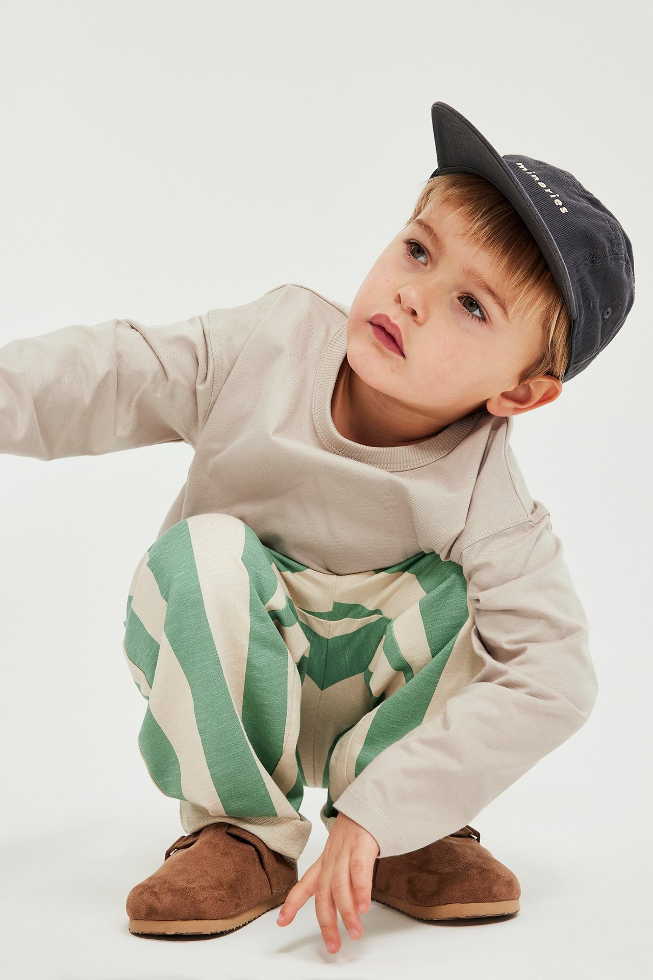Widok z przodu: Dziecko ma na sobie beżową koszulkę z długim rękawem, zielone spodnie w paski, ciemnoszarą czapkę i brązowe buty.