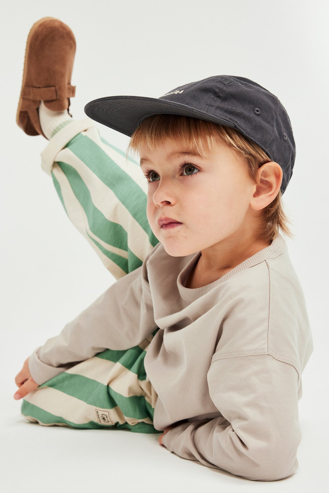 Dziecko w beżowej koszulce z długim rękawem, pasiastych spodniach, ciemnoszarej czapce i brązowych kapciach.