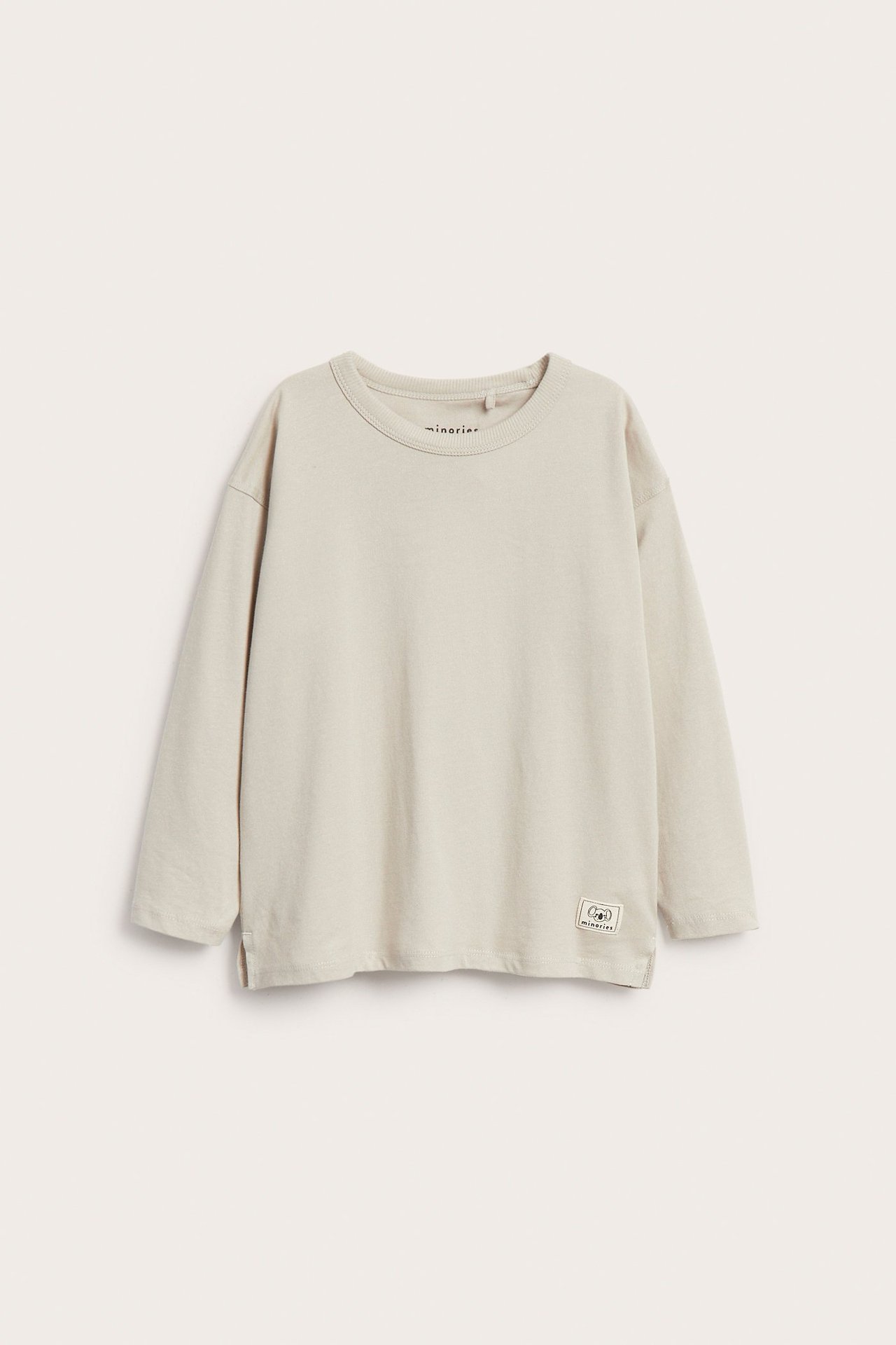 Widok z przodu: beżowa koszulka dziecięca z długim rękawem, okrągłym dekoltem i małym znaczkiem.