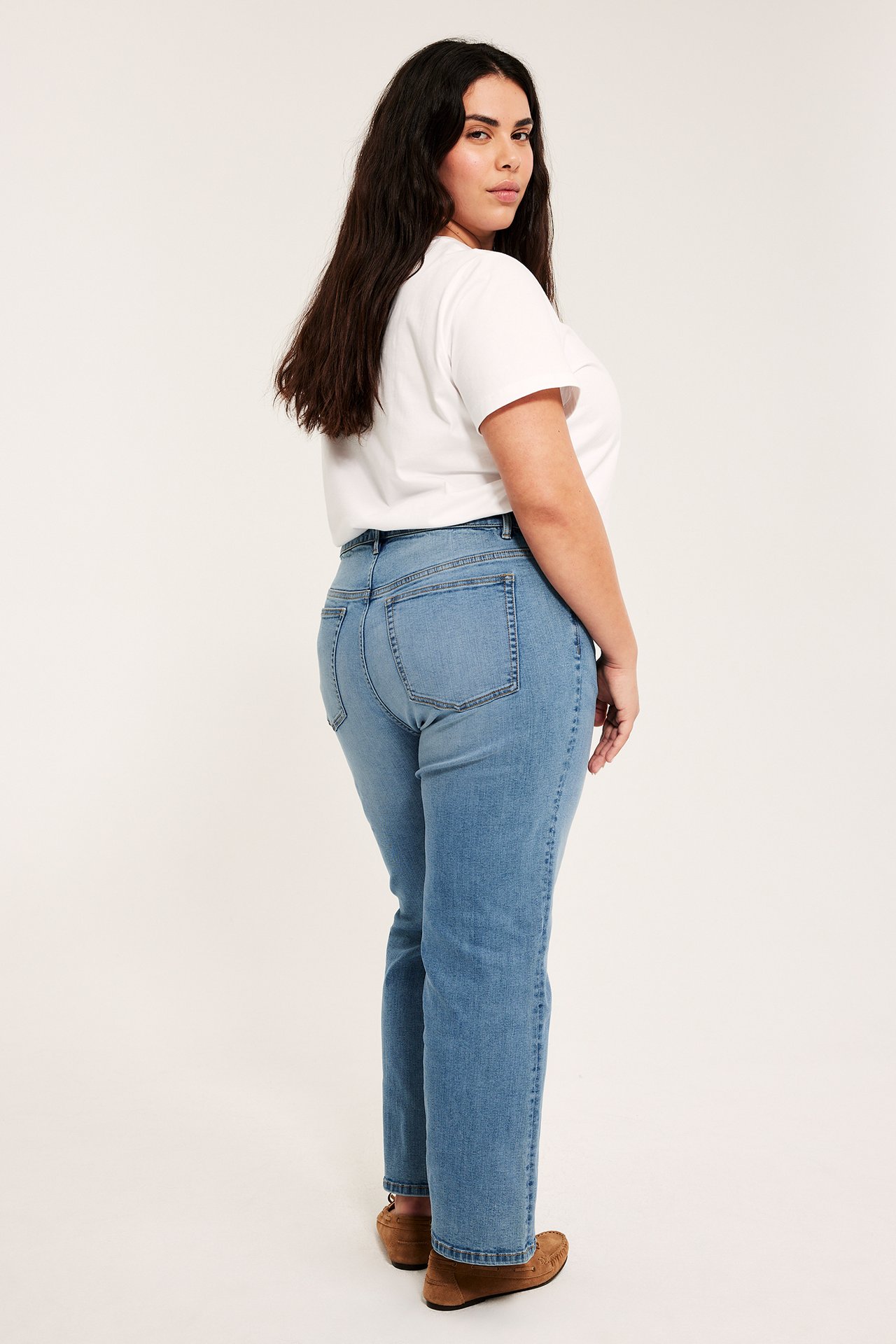 Widok z boku i tyłu kobiety w jasnych dżinsach cropped flare, wysokim stanem, białym t-shircie i brązowych butach.