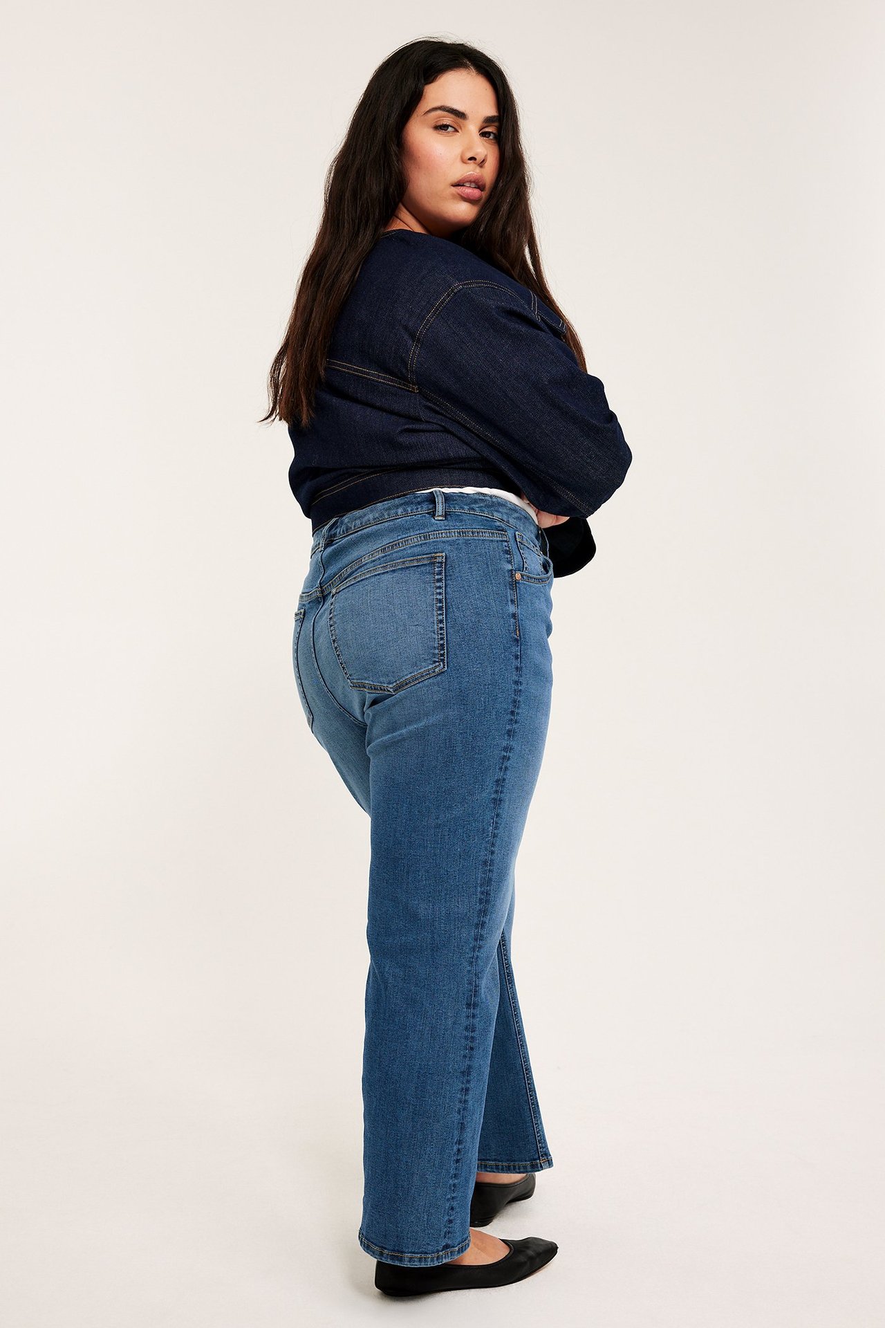 Widok z boku: kobieta w niebieskich dżinsach cropped flare, ciemnej kurtce dżinsowej i czarnych baletkach.