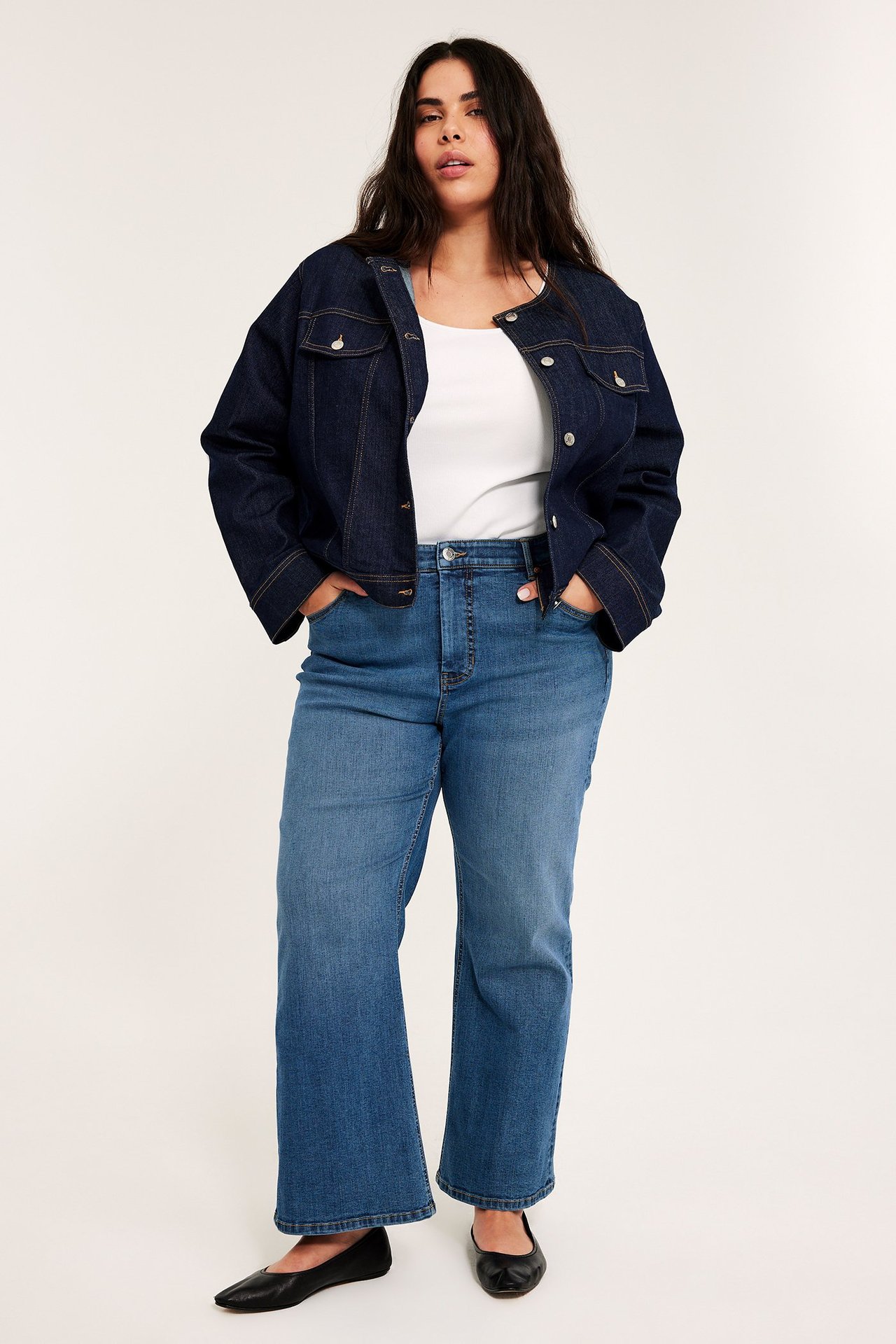 Kvinna i mellanblå högmidjade croppade flare-jeans, vit topp, mörkblå jeansjacka och svarta skor. Framifrån.
