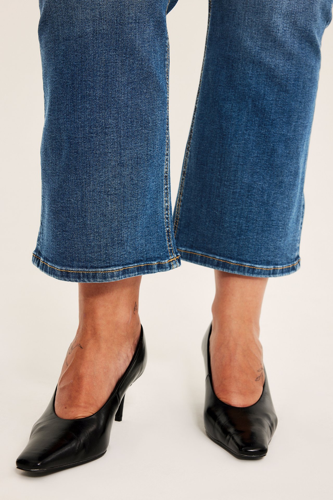 Närbild framifrån på blå crop-jeans i denim med gula sömmar och svarta spetsiga högklackade skor.