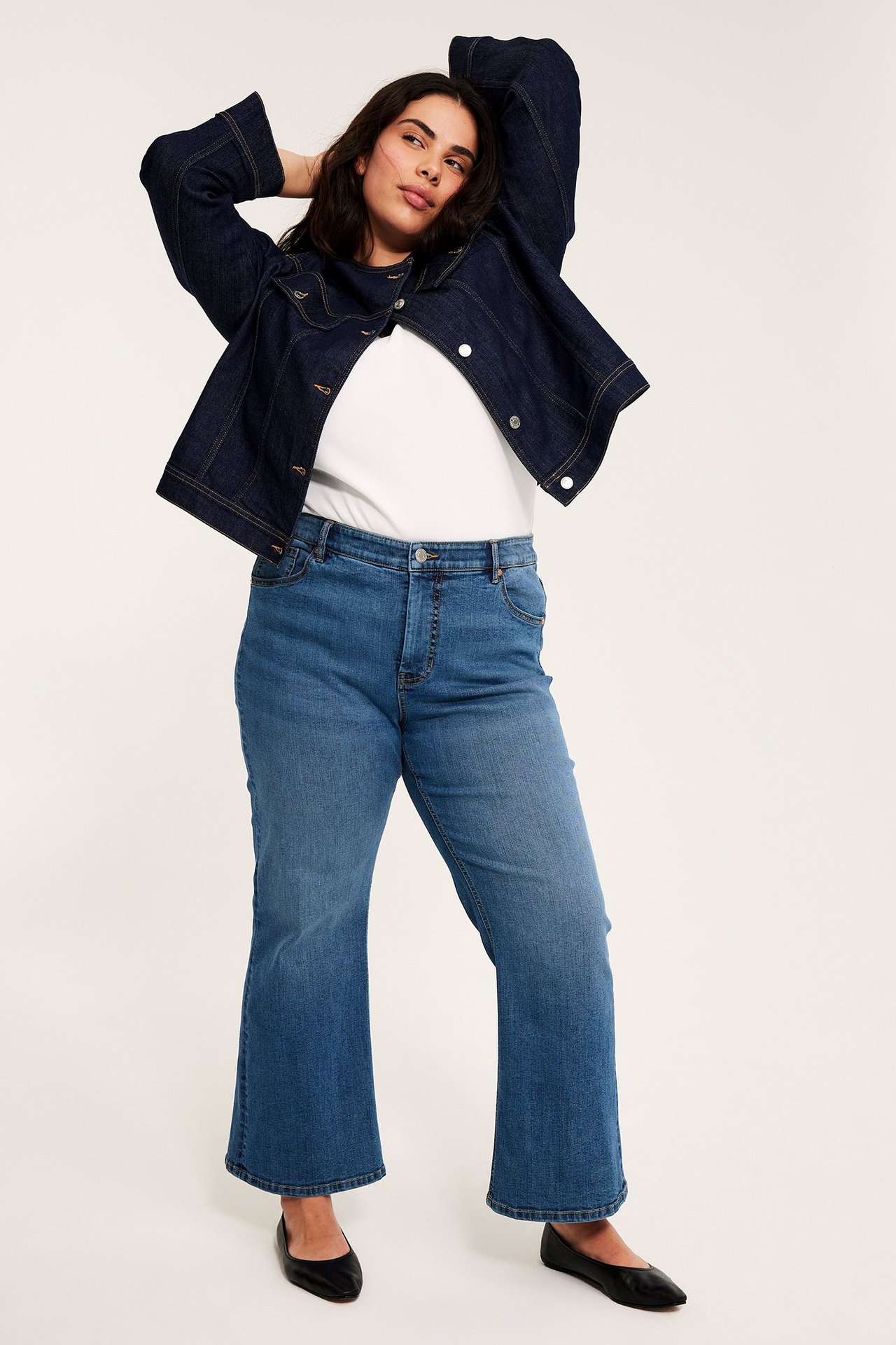 Frontvy av kvinna i blå denim high-waist cropped flare jeans, vit t-shirt, mörkblå jeansjacka och svarta ballerinaskor.