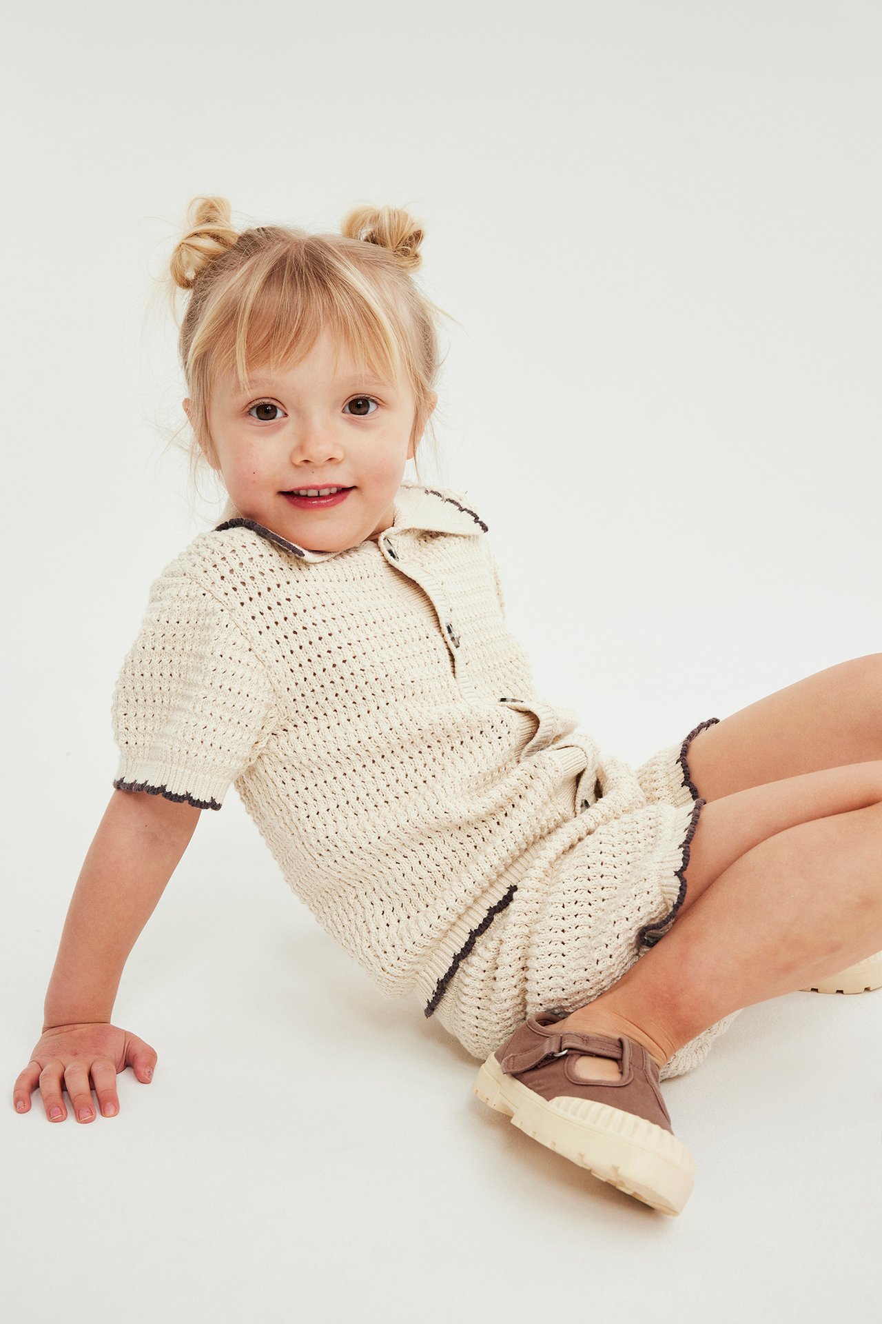 Barn i beige stickat set i bomull; topp och shorts med mörk kant, och bruna skor.