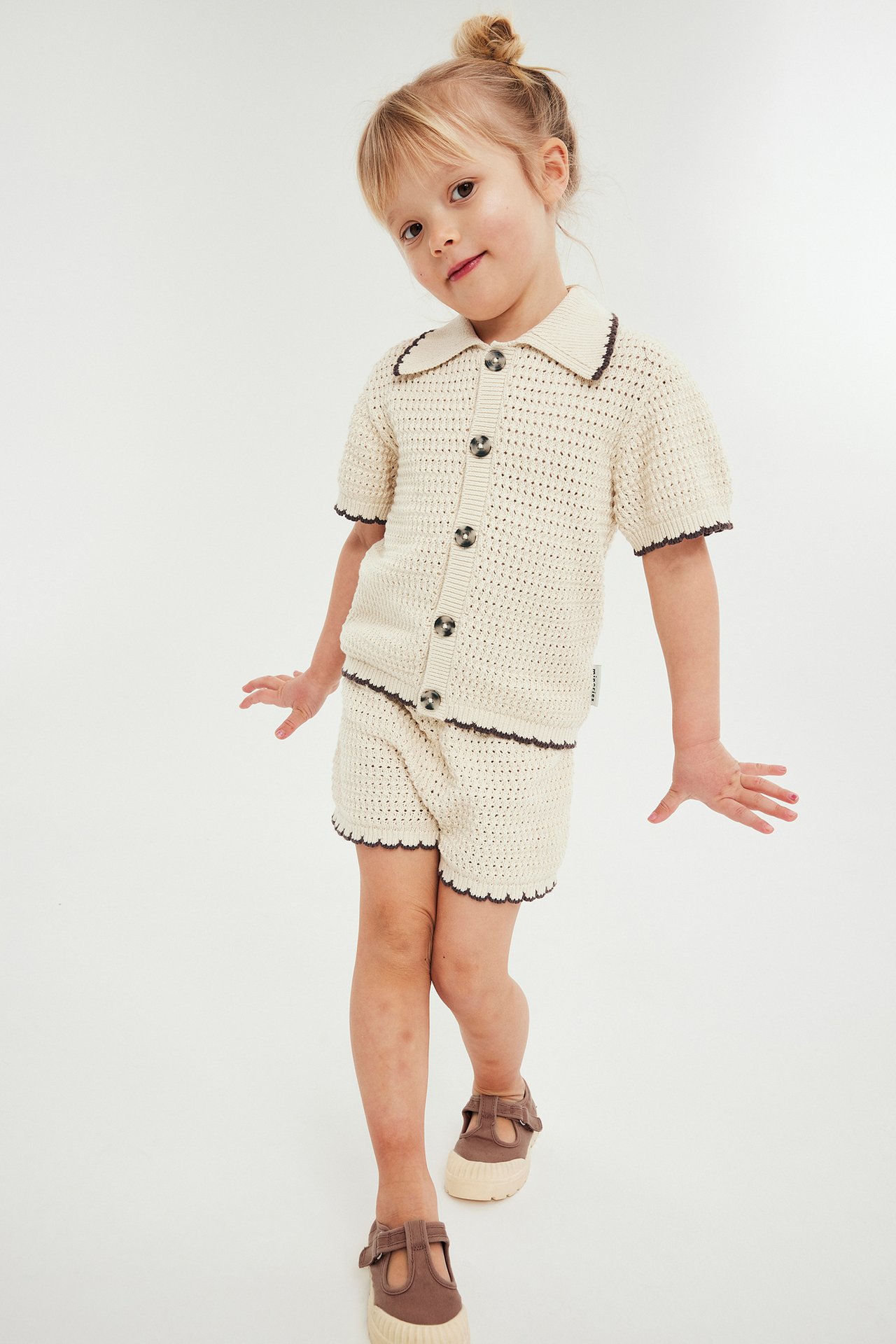 Framsida: Ett barn i beige stickad kortärmad topp med knappar och matchande shorts med vågig kant.