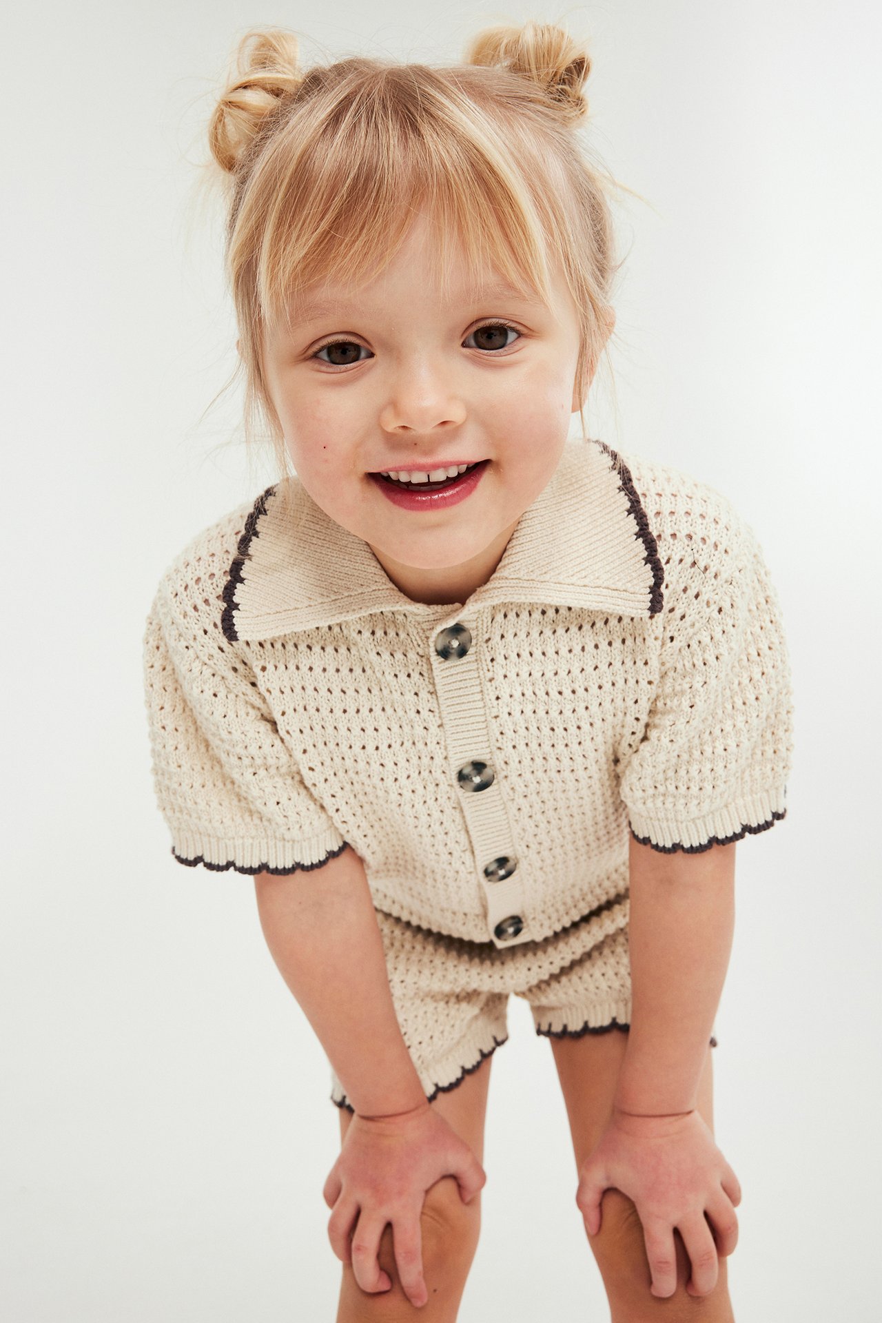Framifrån: Ett leende barn bär beiget stickat set; knapptopp och matchande shorts med mörk kant.