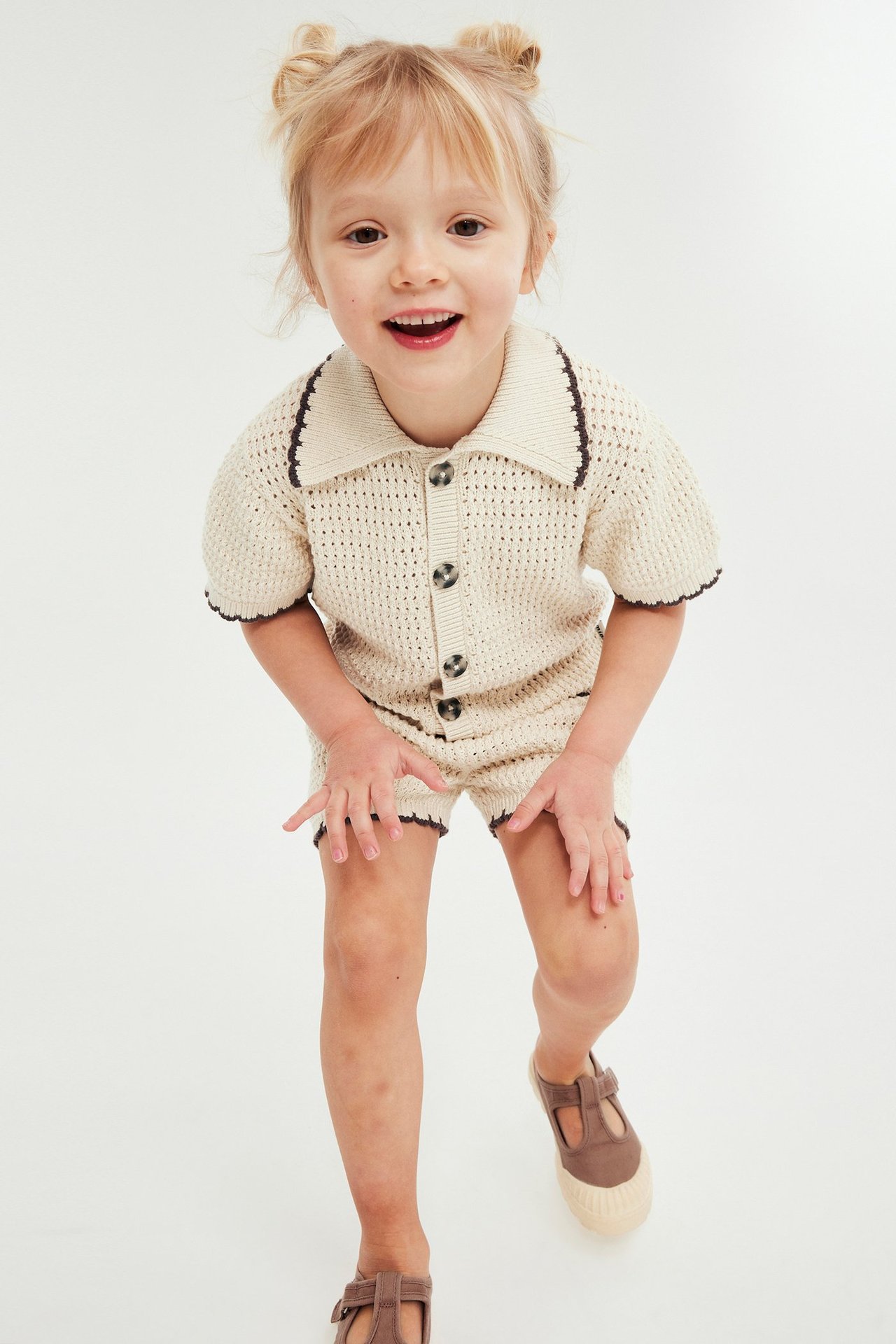 Forfra, et barn i et beige strikket sett med kragegenser og shorts, samt brune sko.