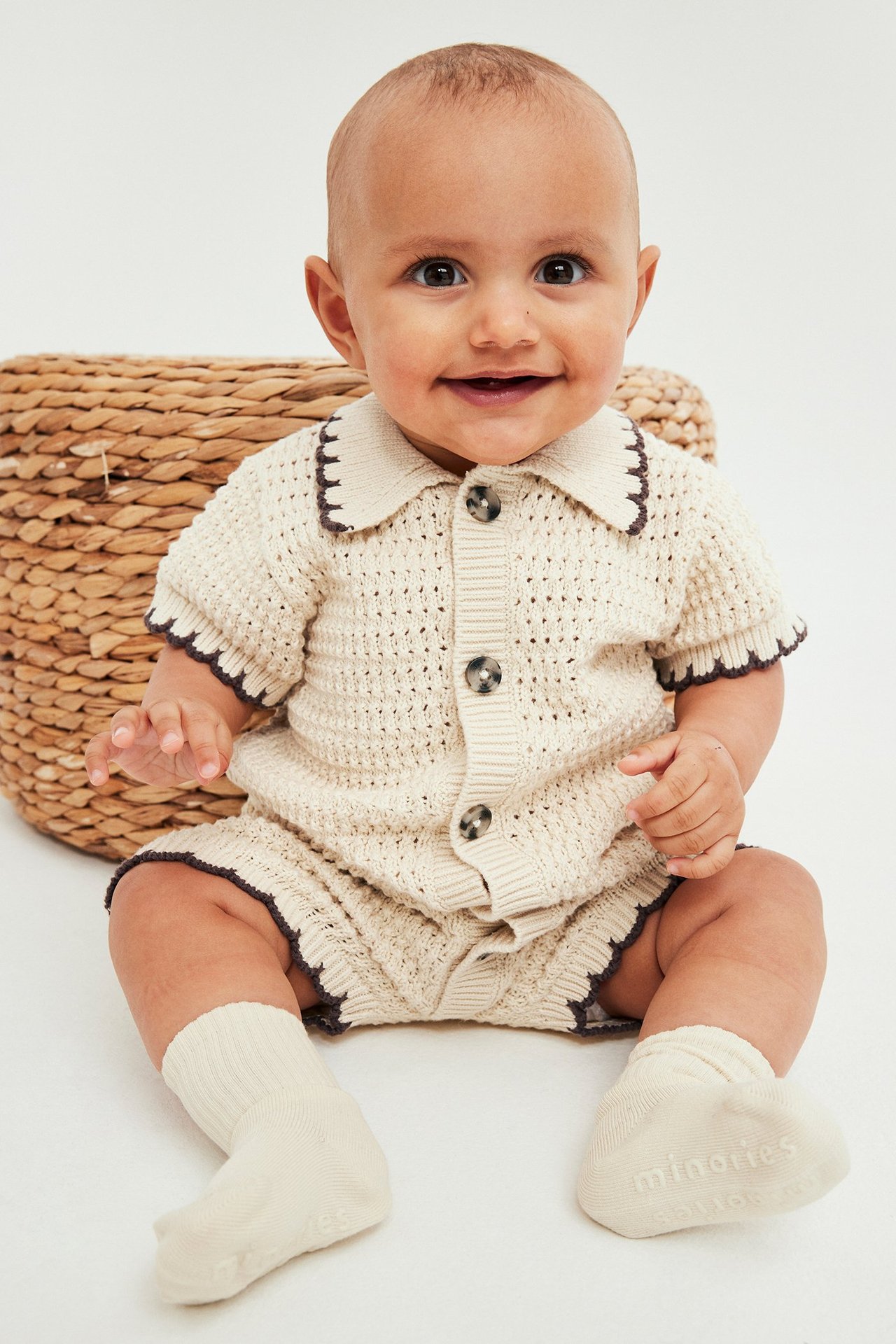 Framsida: Baby i beige stickad kortärmad body med brun kant på krage, knappar och beige strumpor.