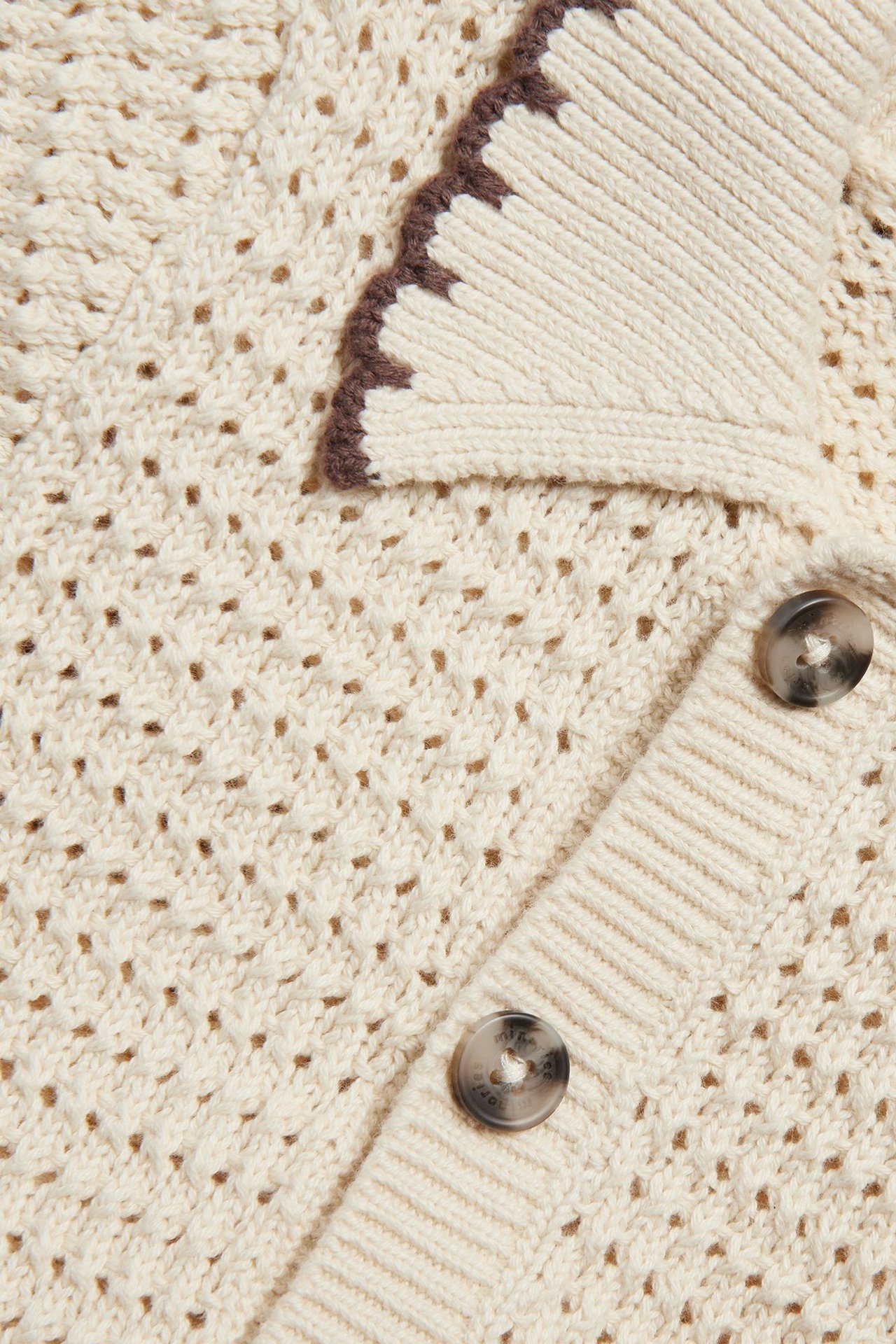 Närbild: Beige stickad babybody med hålmönster, brun kantad krage och knappar fram.
