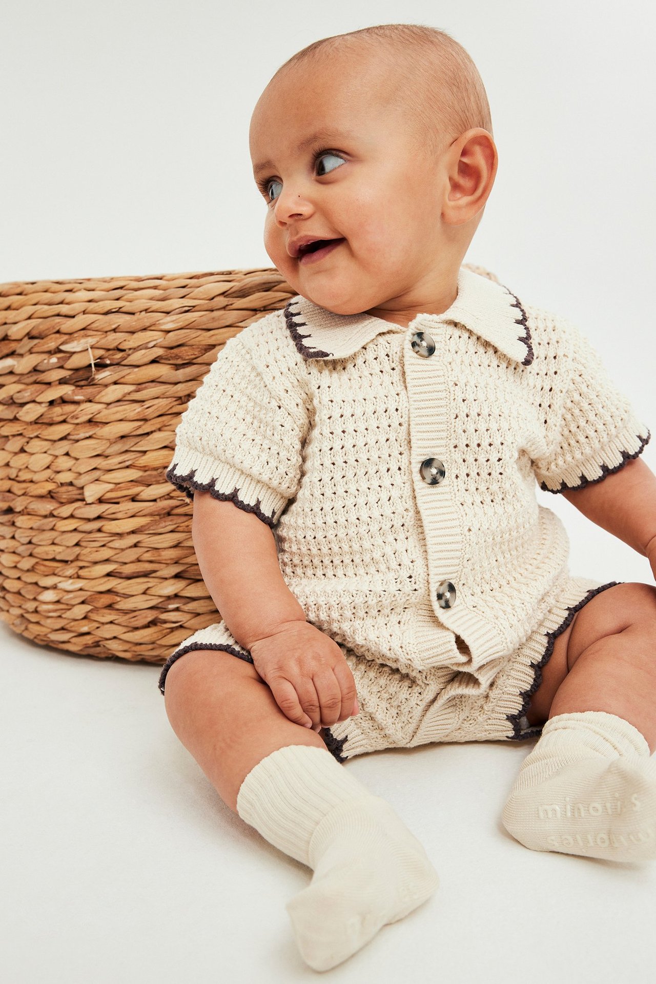 Frontvy: Baby i beige stickad bomullsbody med krage, knappar, mörk kant, och ljusbeige strumpor.