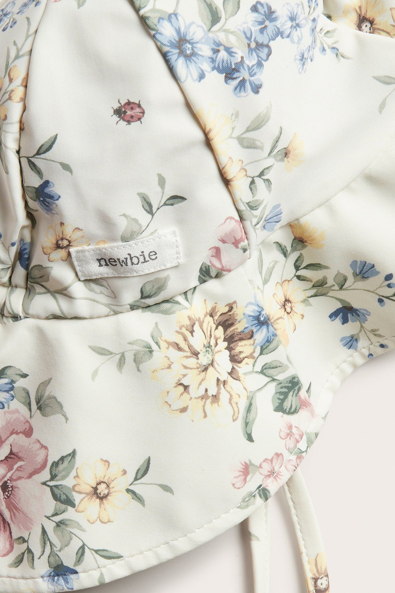 Nærbilde av en offwhite blomstret solhatt med knytebånd og en Newbie-etikett.