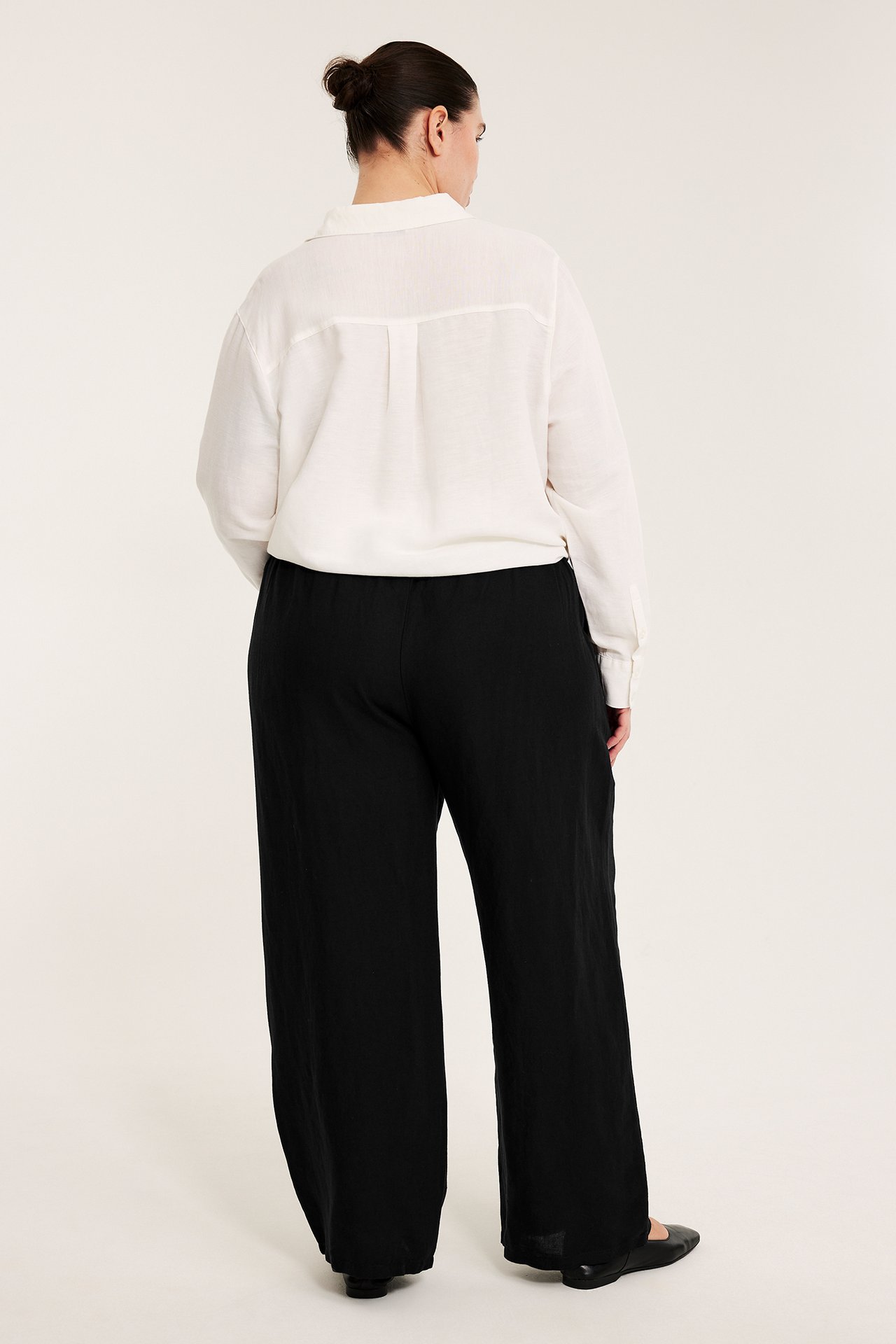 Widok z tyłu kobiety w czarnych spodniach z lnu z marszczonym pasem i białej koszuli.