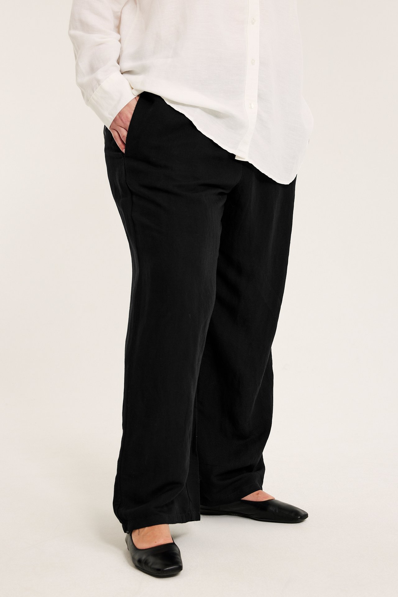 Widok z przodu kobiety w czarnych spodniach z mieszanki lnu ze ściągaczem, białej koszuli i czarnych balerinach.