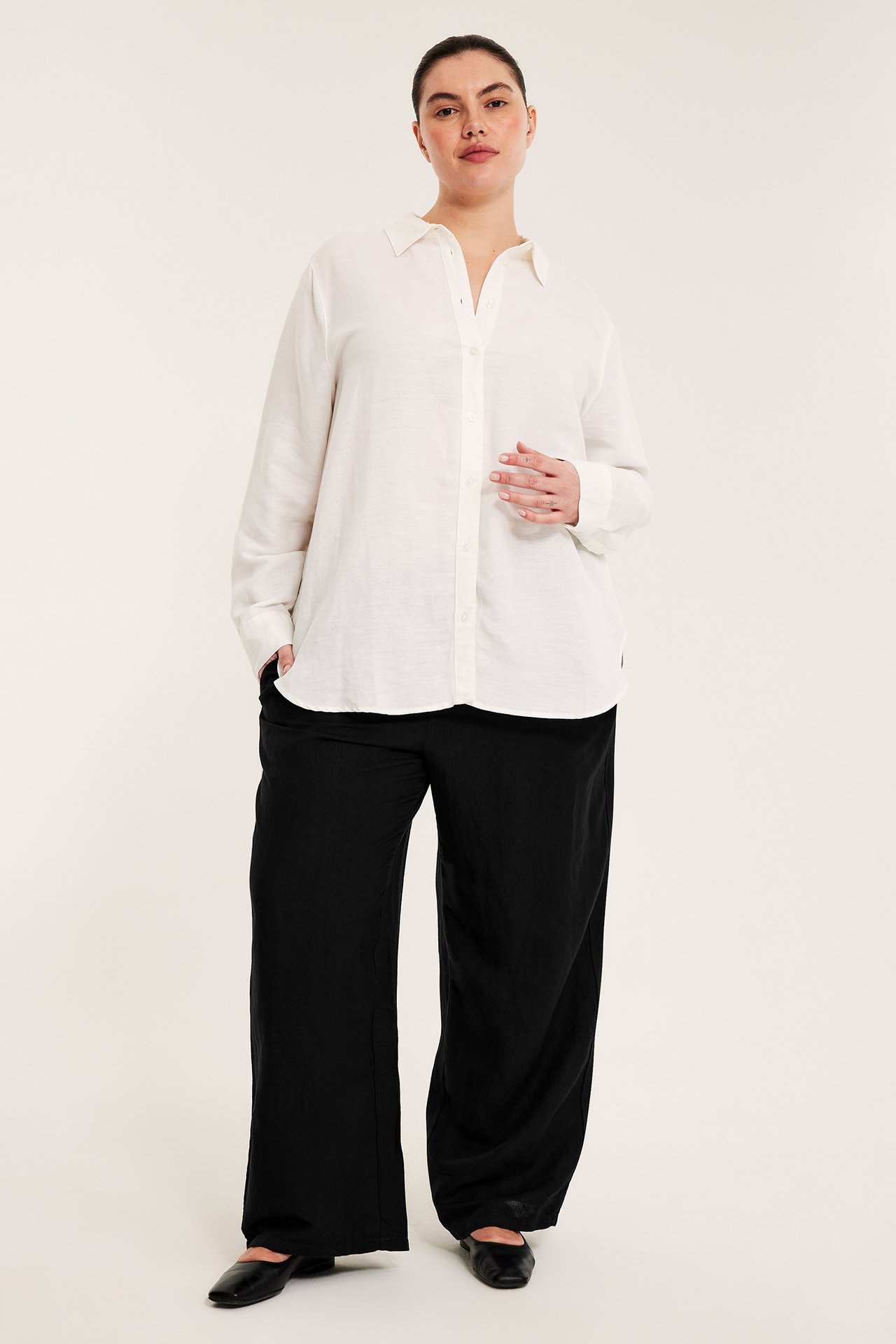 Widok z przodu: Kobieta w czarnych szerokich spodniach z lnu, białej lnianej koszuli i czarnych balerinach.