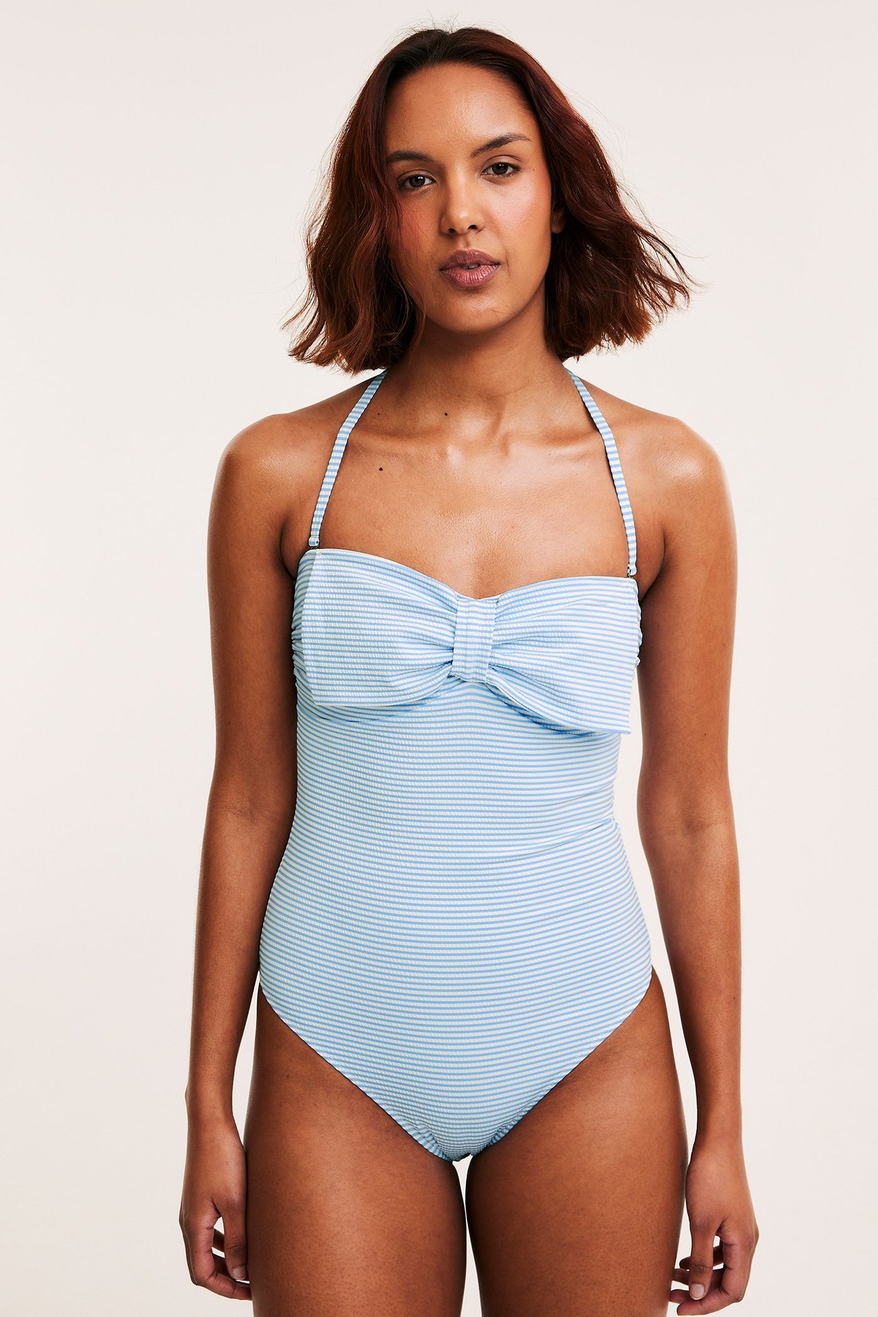 Nainen edestäpäin, yllään raidallinen vaaleansinivalkoinen uimapuku rusetilla ja halterneckilla.