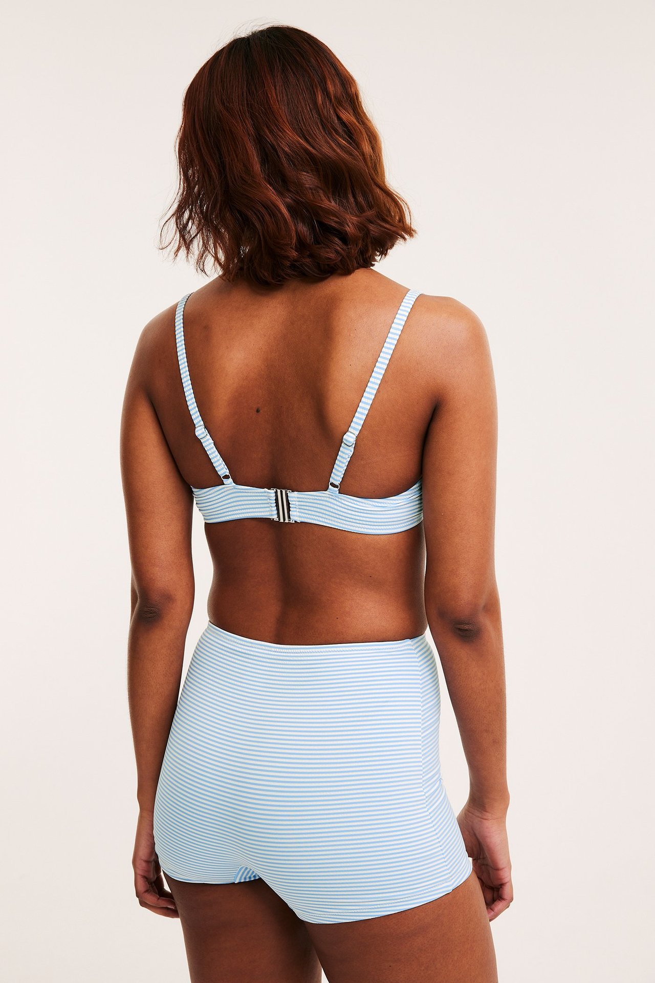 Ryggvy av en kvinna i ljusblå- och vitrandig högmidjad bikinitrosa i shortsmodell och matchande bikiniöverdel.