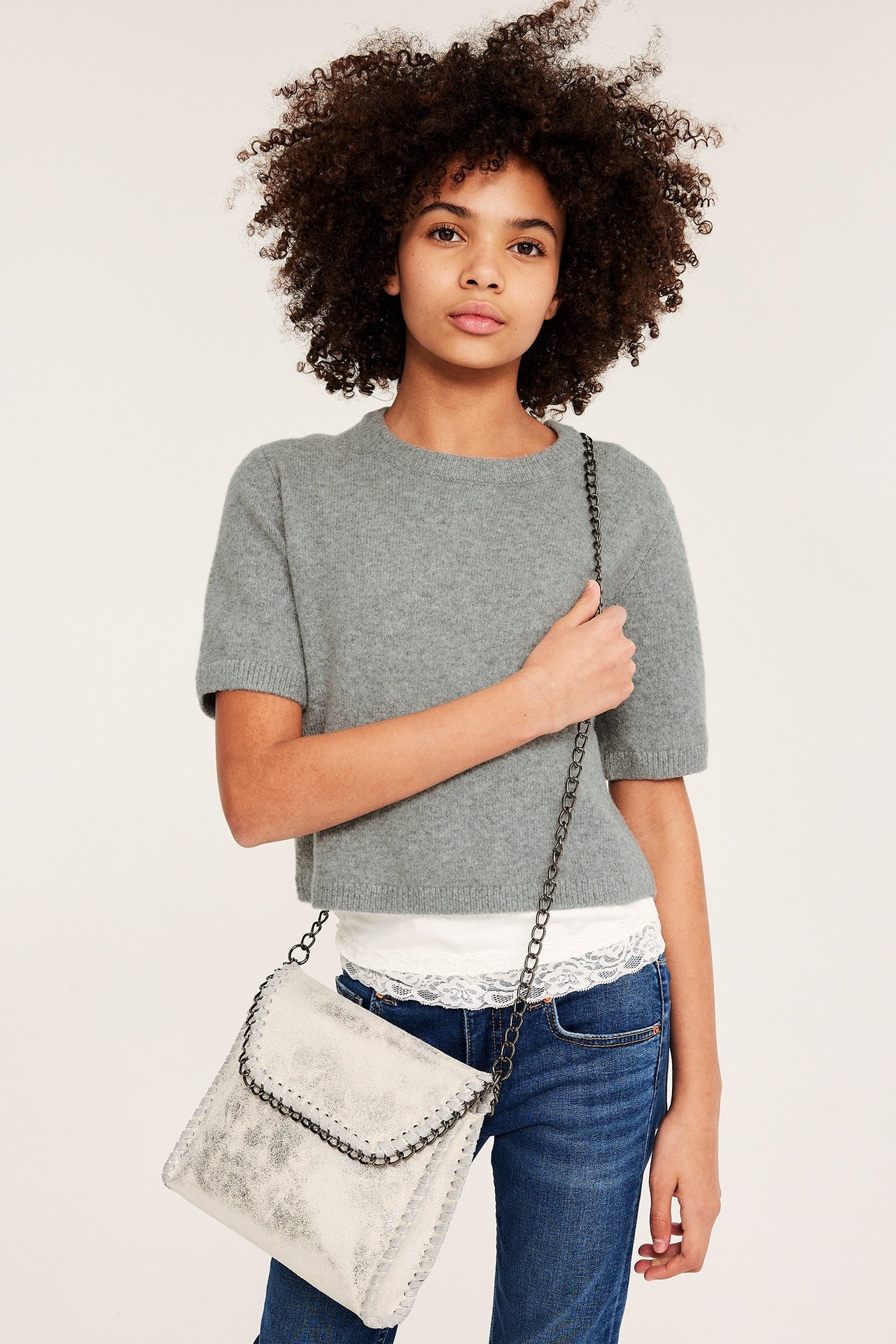 Jente i lys grå finstrikket kortermet genser, hvit blonde topp, blå jeans, holder sølvveske.