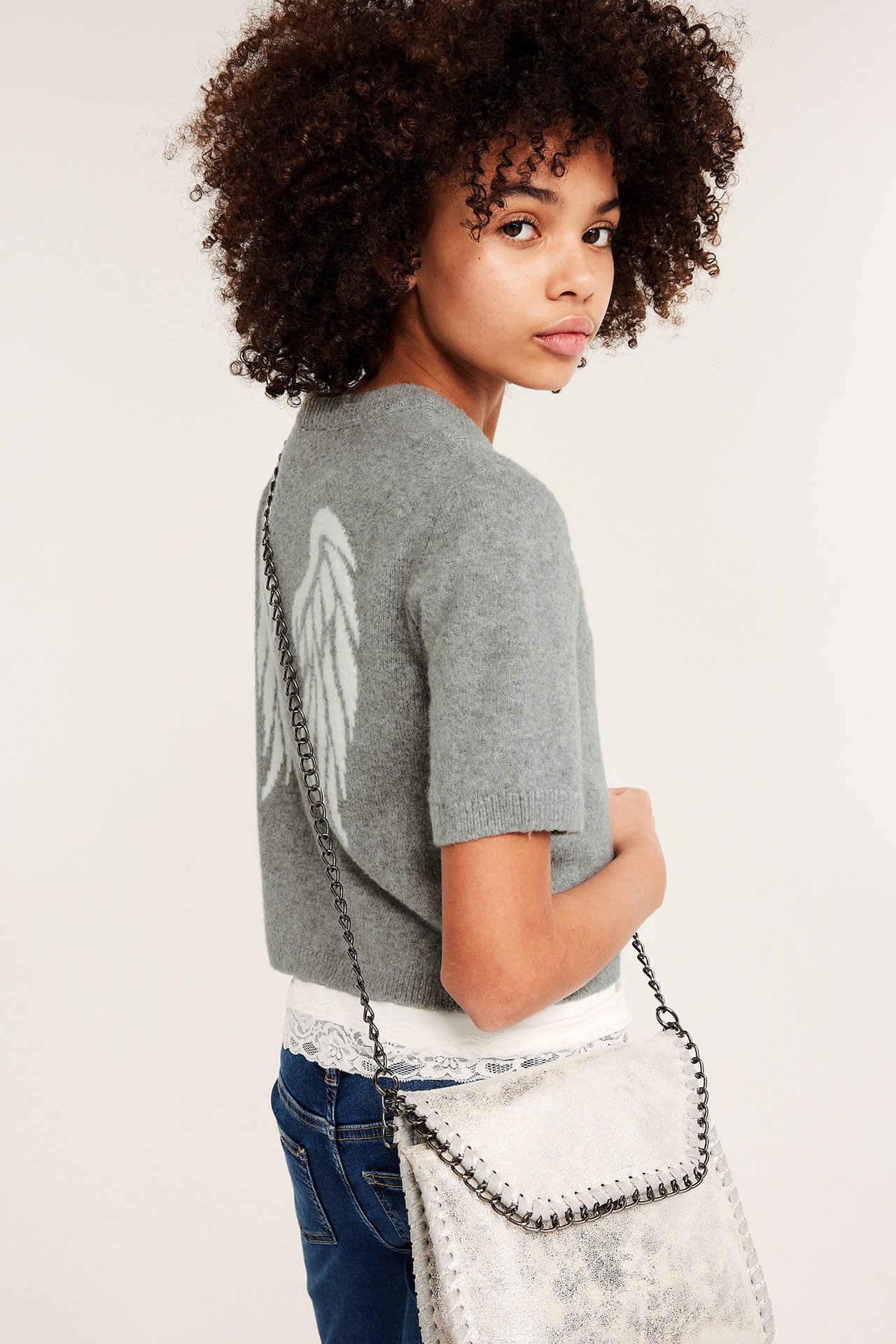 Sida vy av ett barn i ljusgrå finstickad t-shirt med vita vingar, vit topp, blå jeans och silverväska.