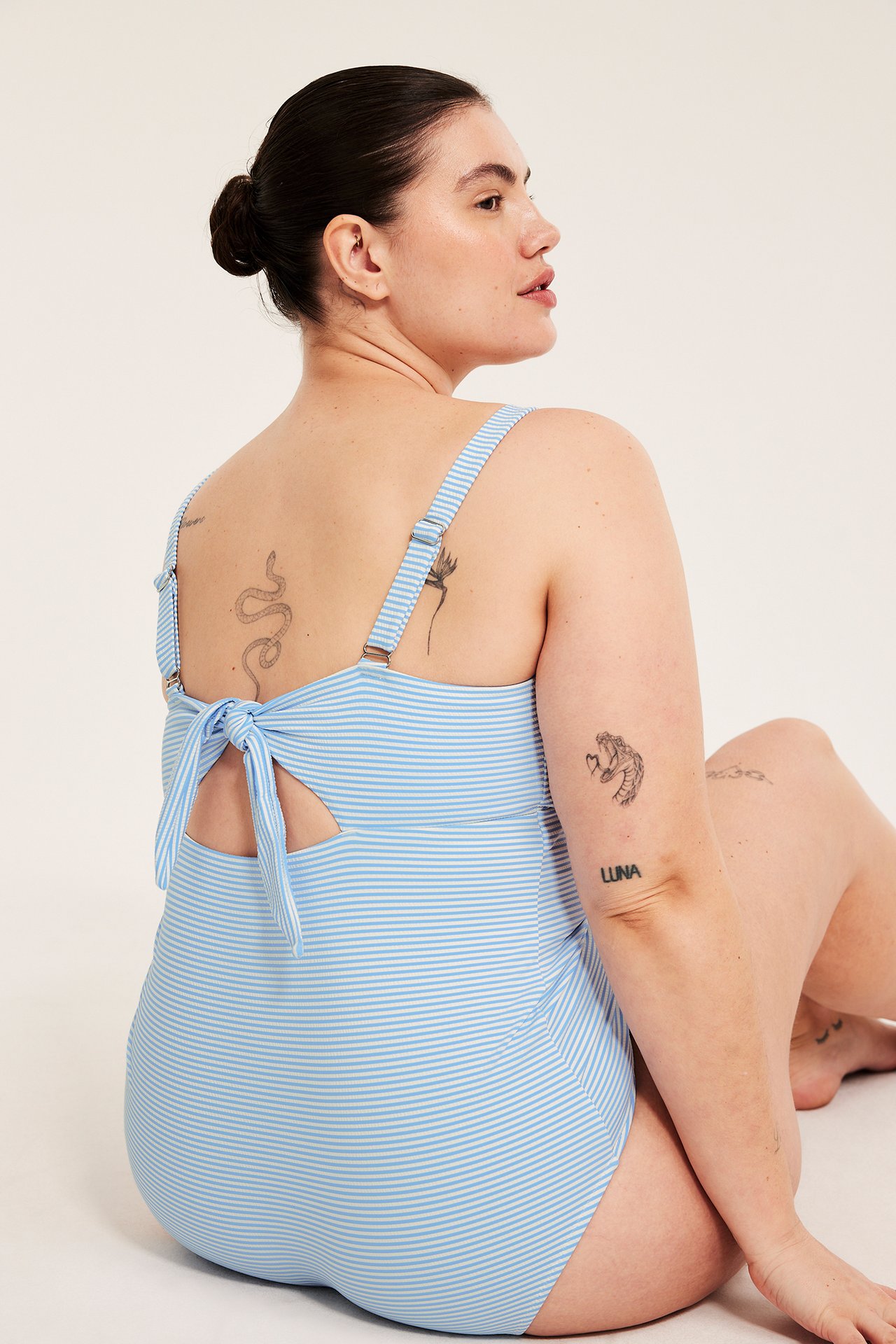 Naisen takakuva vaaleansinisessä raitaisessa uimapuvussa, jossa säädettävät olkaimet ja solmittava selkäosa.