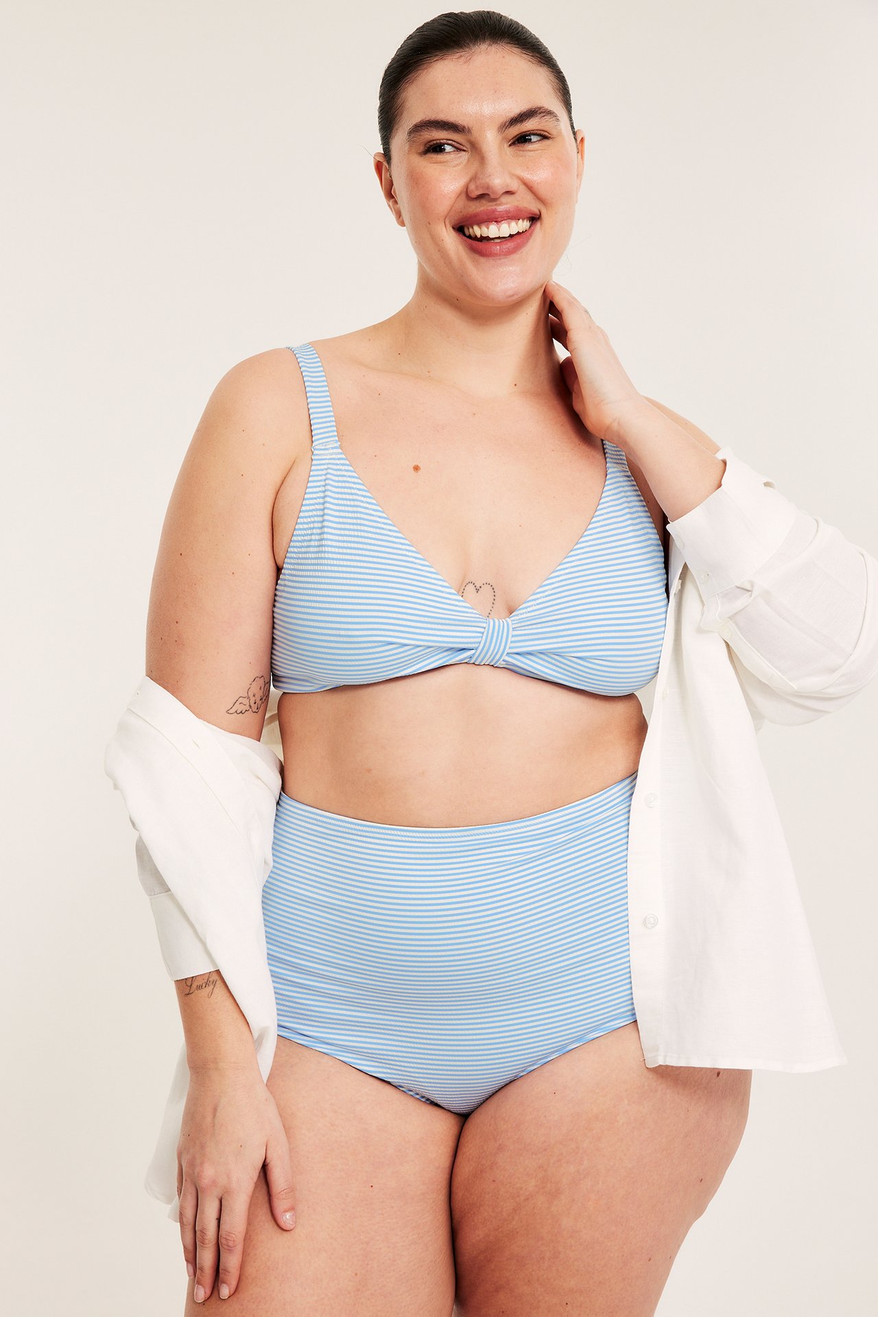 Edestä, hymyilevä nainen vaaleansinisissä raidallisissa korkeavyötäröisissä bikinin alaosissa, vastaava yläosa ja valkoinen paita.