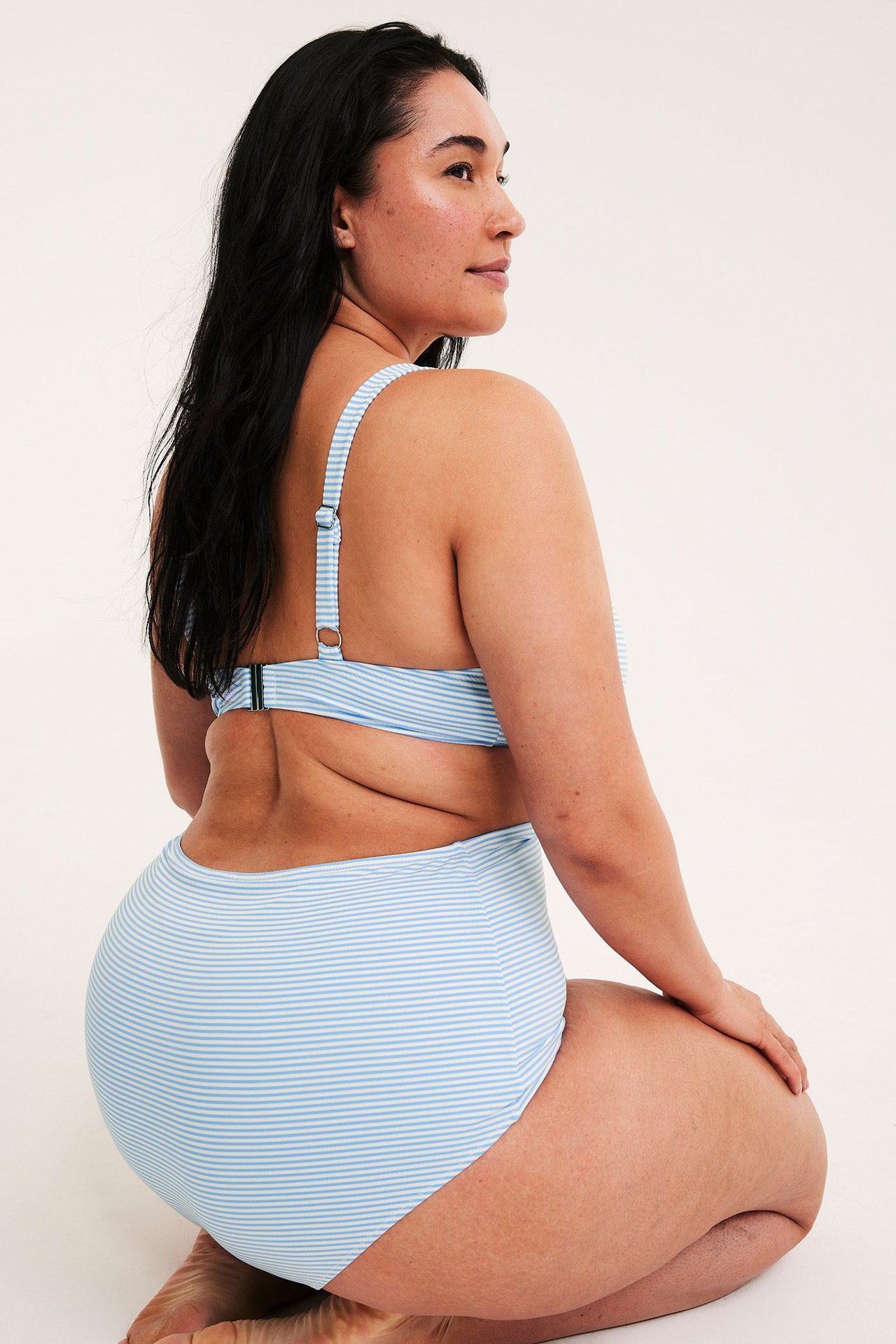 Side-bakfra bilde av kvinne knelende i lyseblå stripet høy midje bikinitruse og matchende topp.