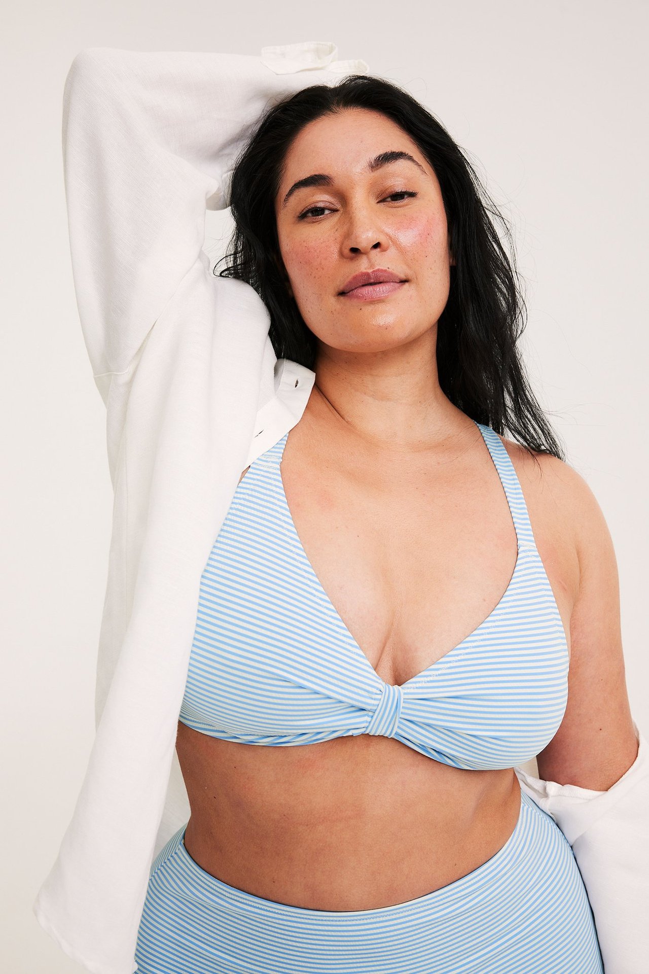 Edestä: nainen yllään vaaleansininen raidallinen bikinitoppi solmittu edestä, yhteensopivat alaosat ja valkoinen avoin paita.